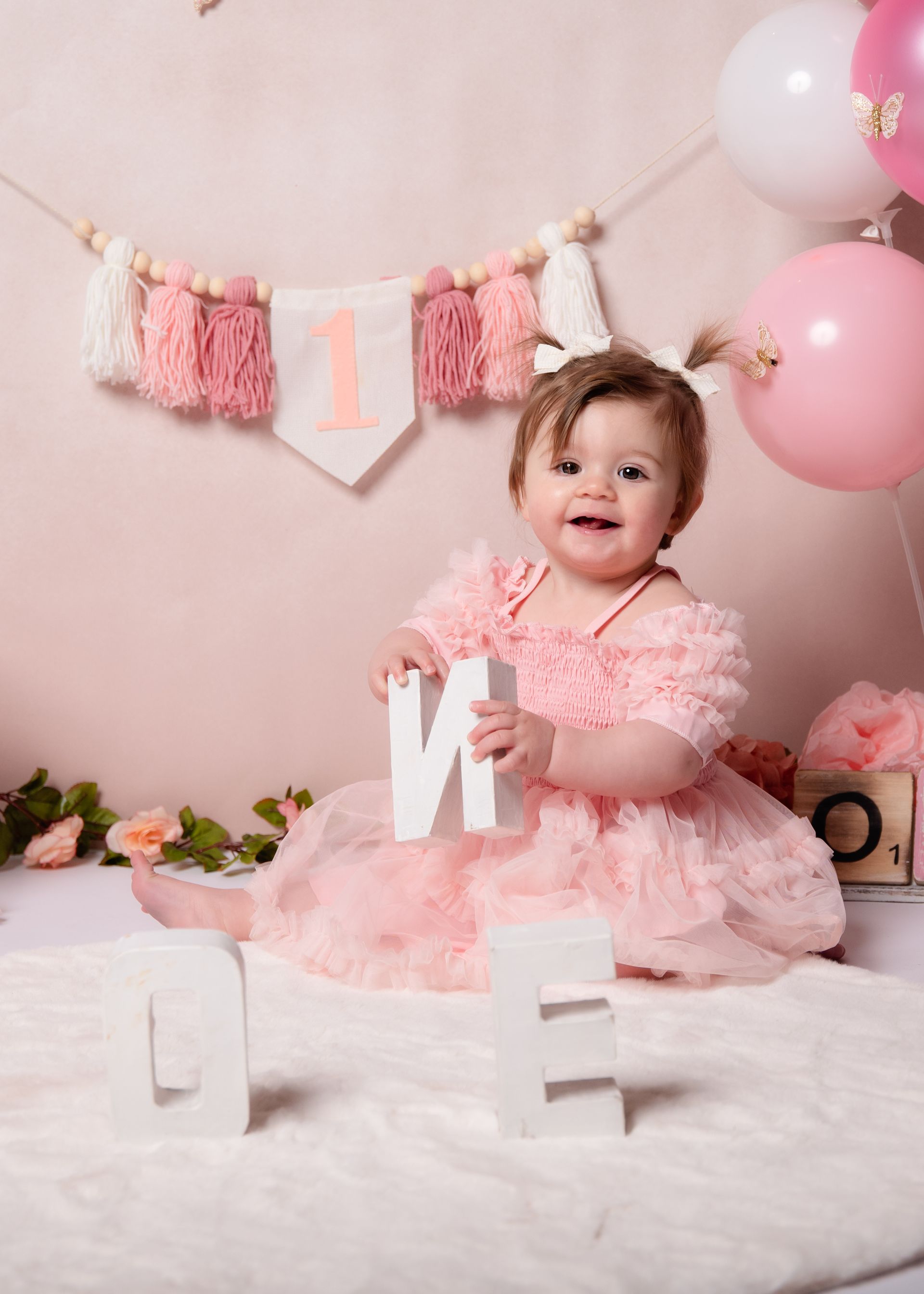 Een babymeisje in een roze jurk zit op een bed met de letter m in haar handen.