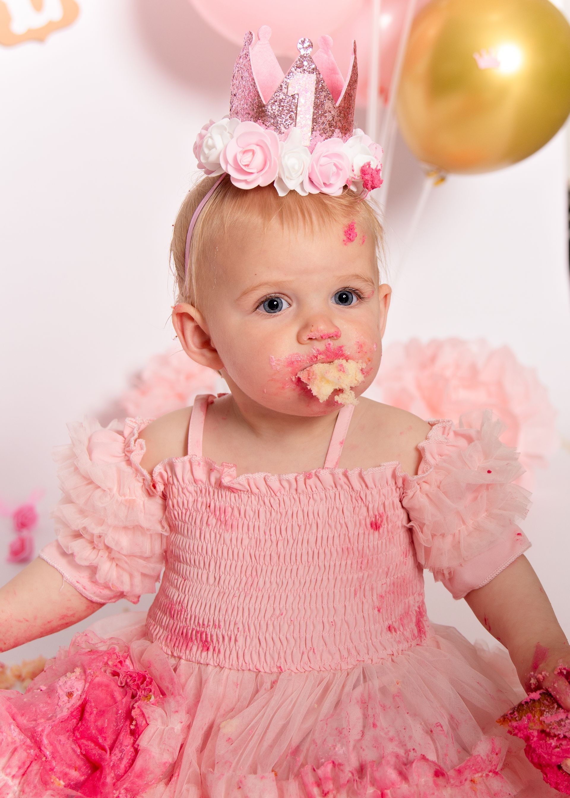 Een babymeisje in een roze jurk en kroontje eet een cupcake.