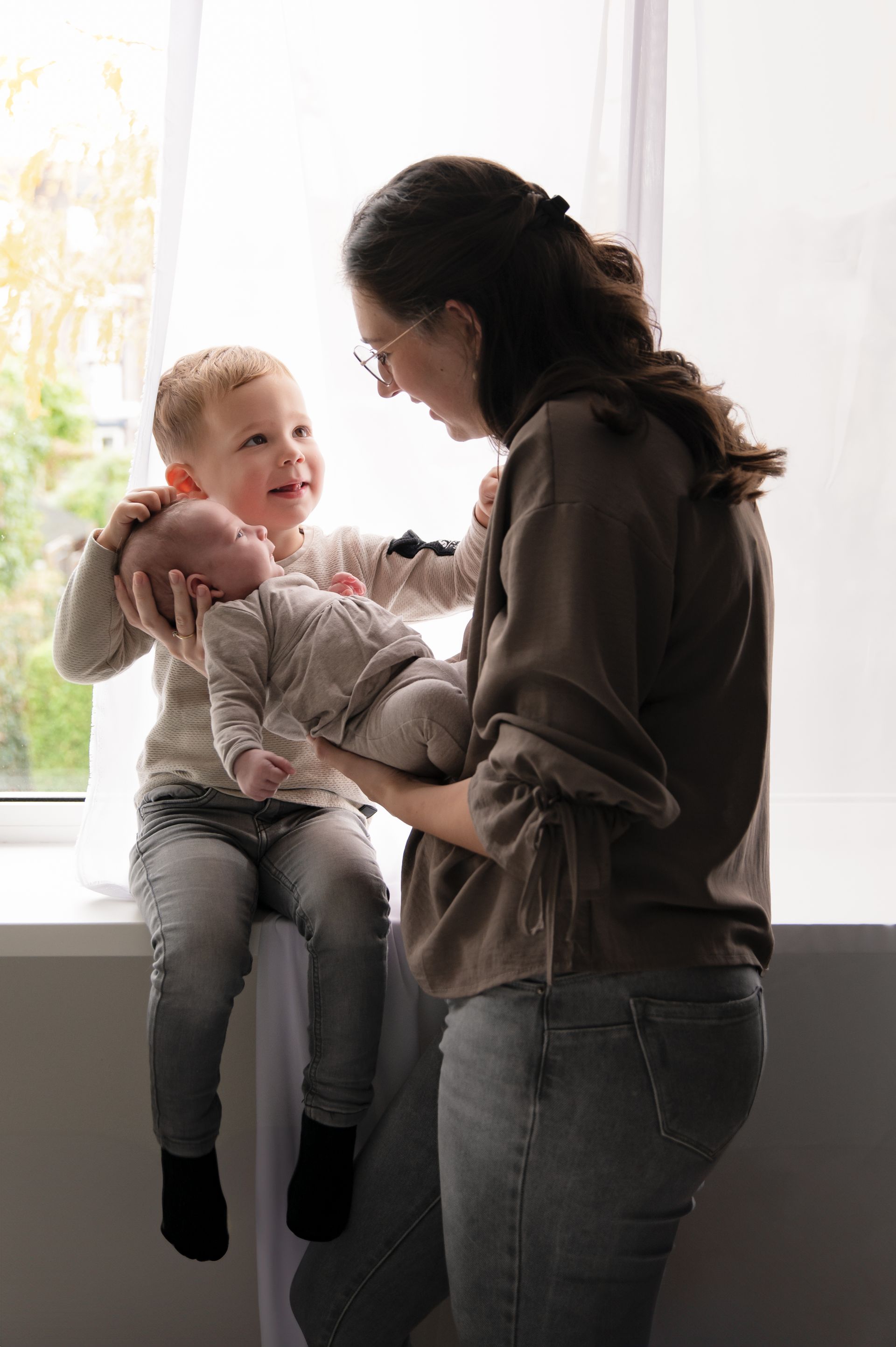 Een vrouw houdt een baby vast en een jongetje zit op de vensterbank.