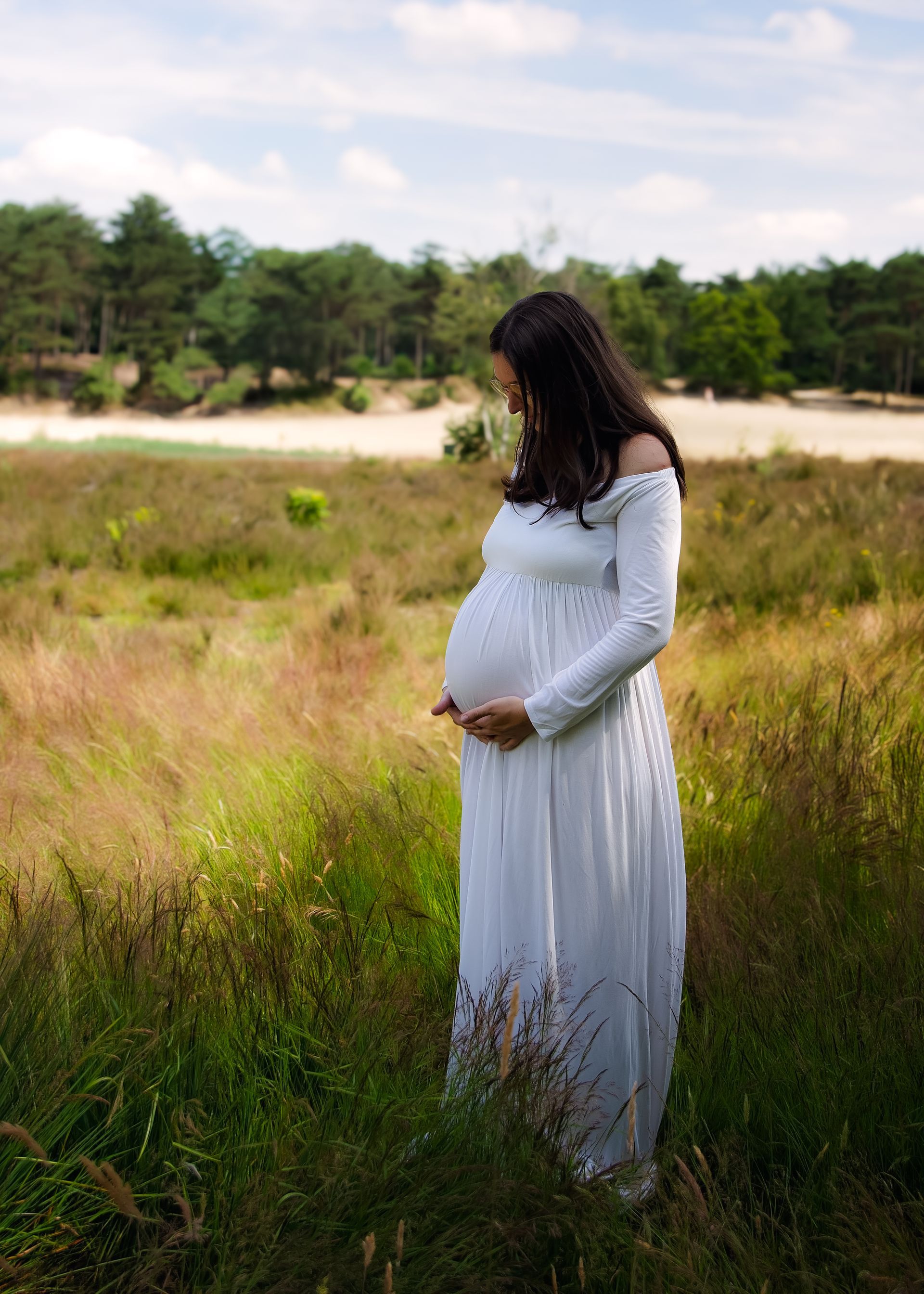 Een zwangere vrouw in een witte jurk staat in een veld en houdt haar buik vast.