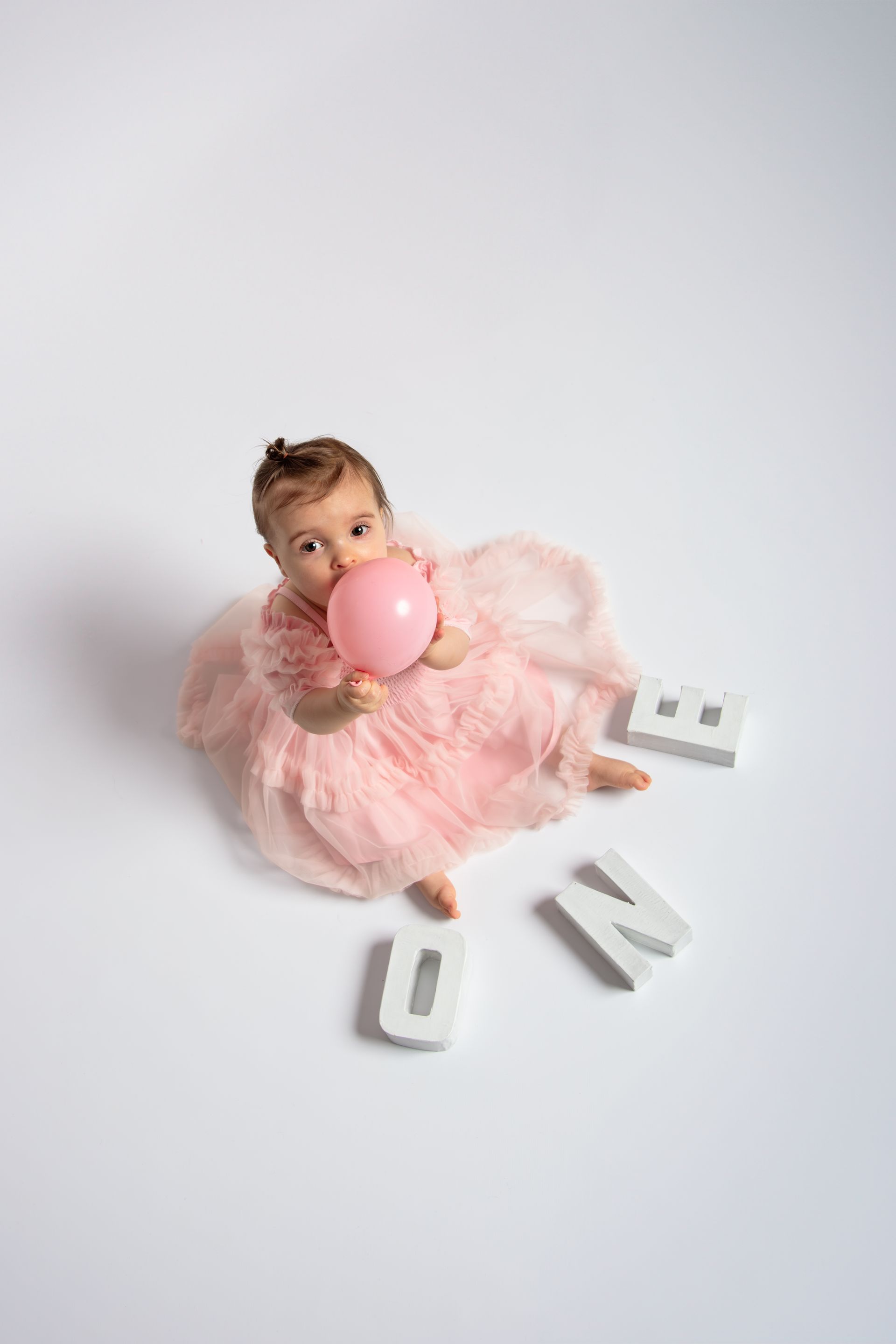Een babymeisje in een roze jurk zit op de grond met een roze ballon in haar handen.