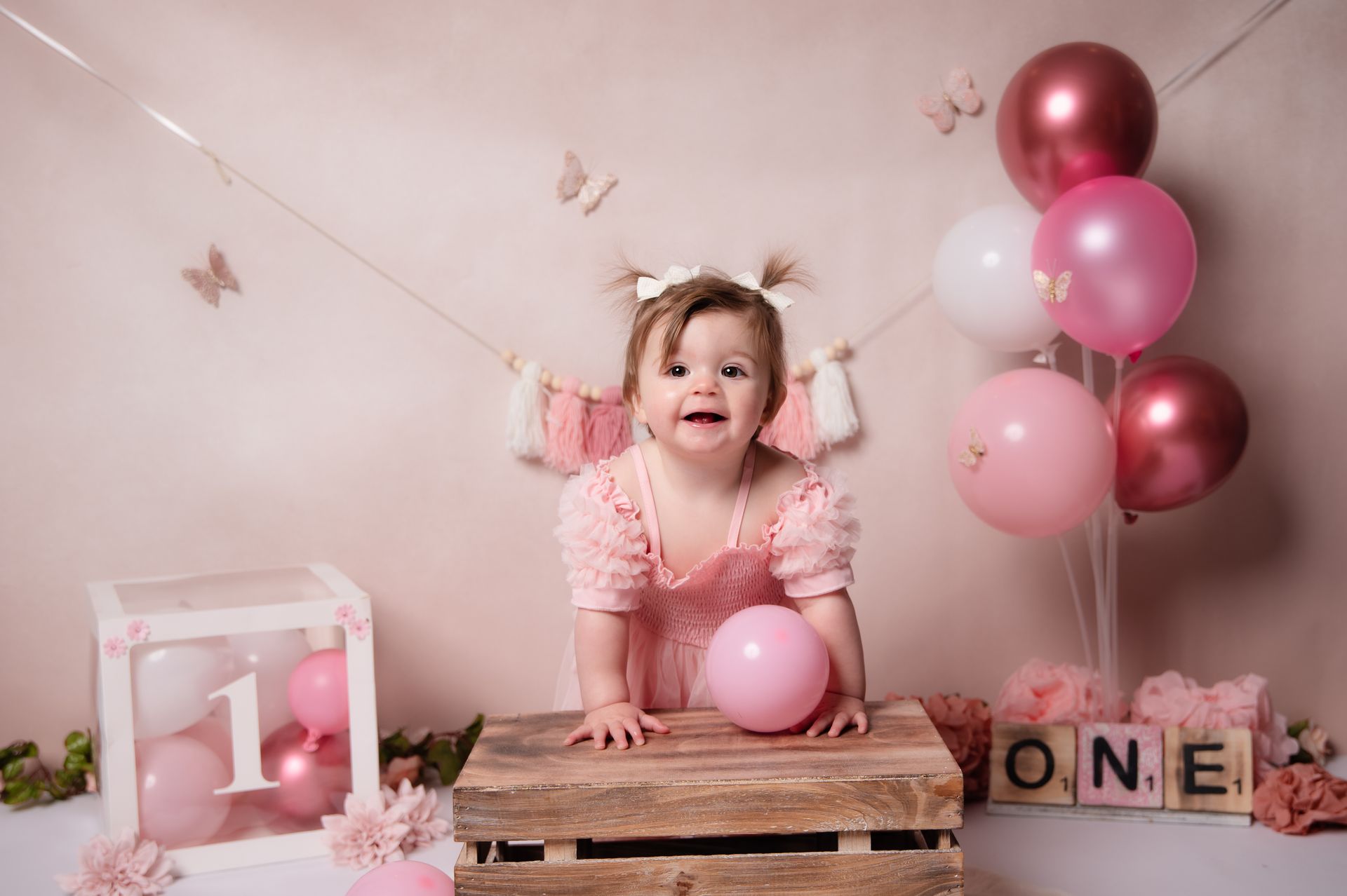Een babymeisje zit op een houten kist met roze ballonnen.