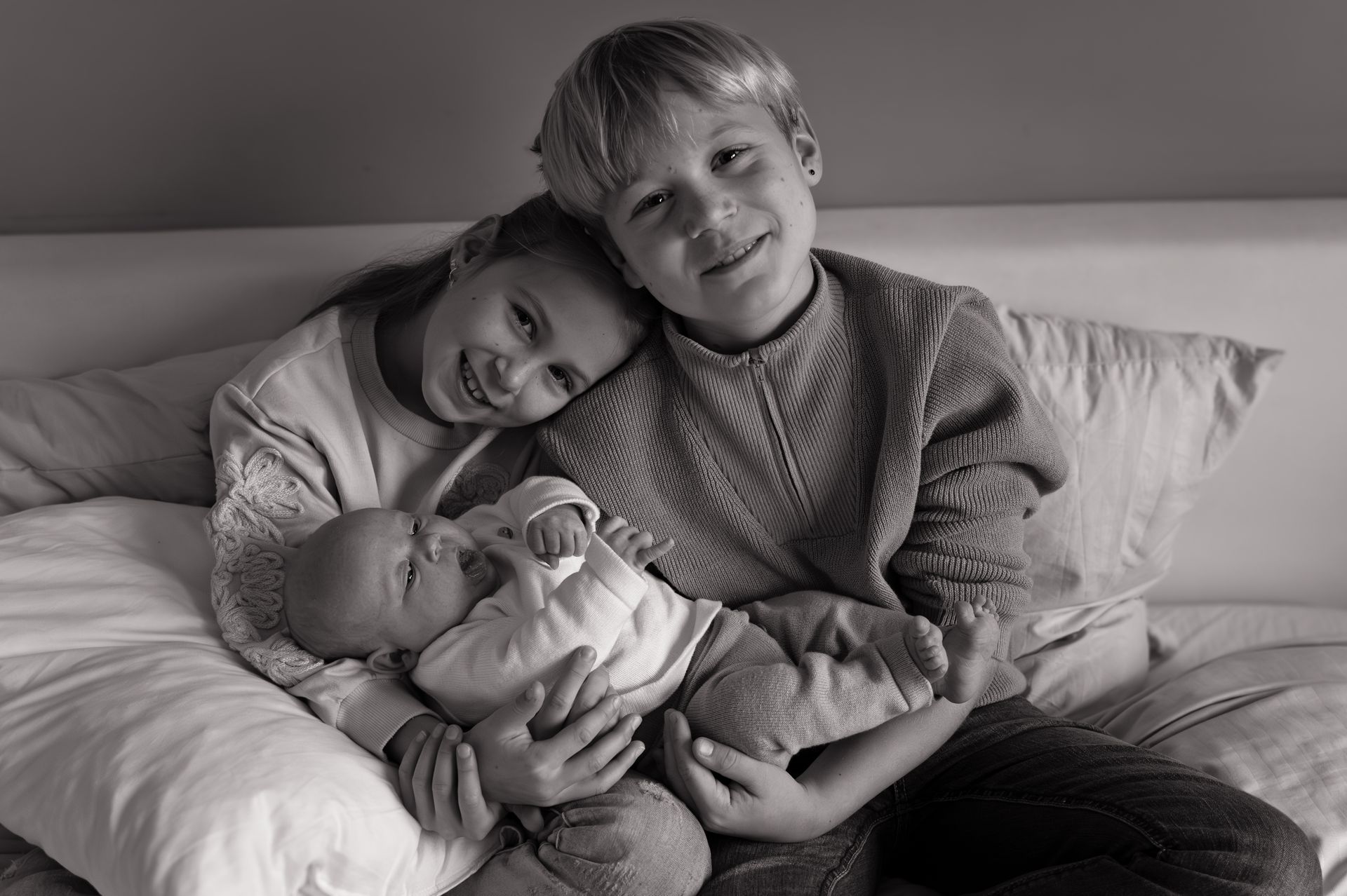 Twee jongens en een meisje zitten op een bed met een baby in hun armen.