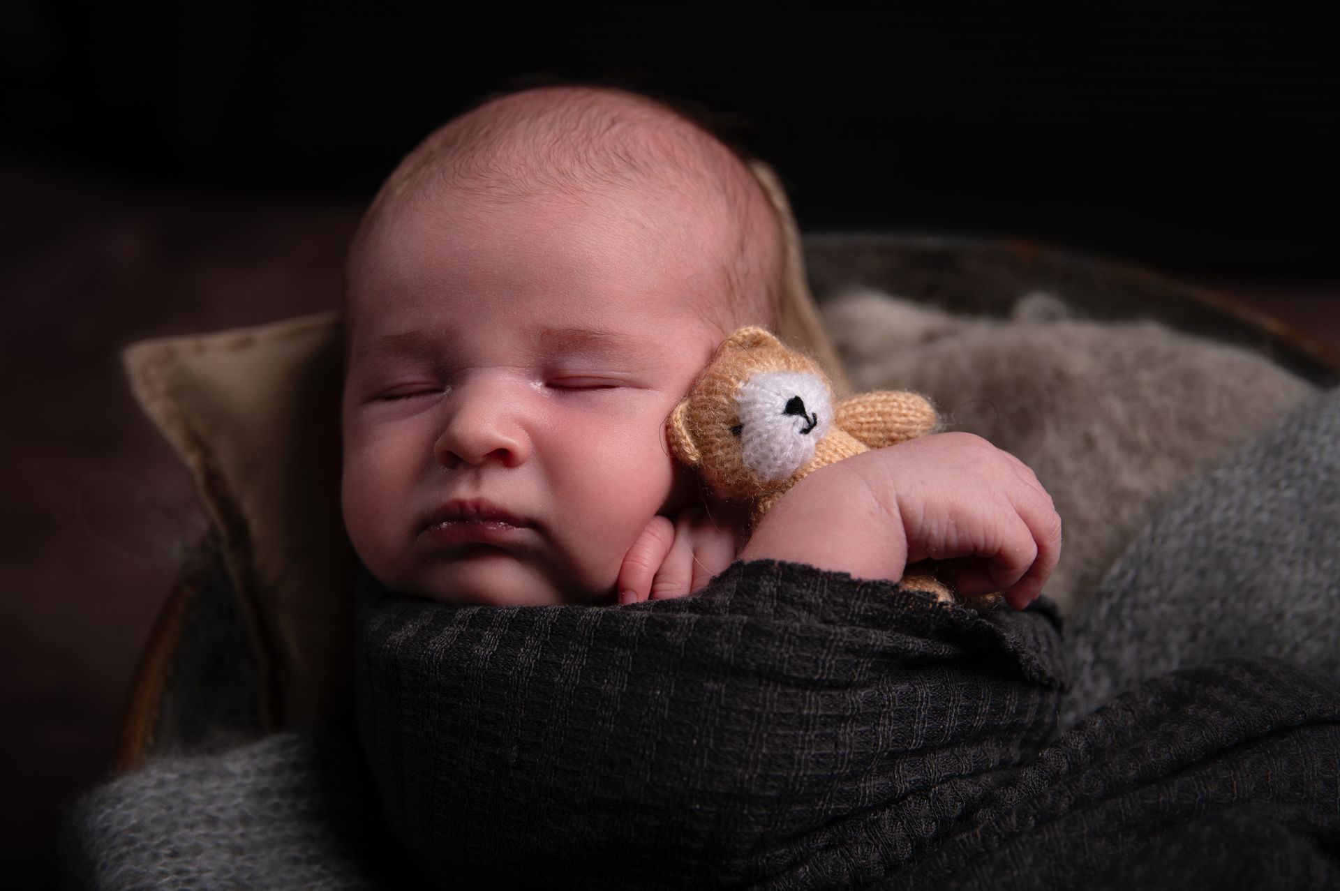 Een pasgeboren baby slaapt in een nest, gewikkeld in grijze dekens en met een kleine bruine teddybeer in zijn armen.