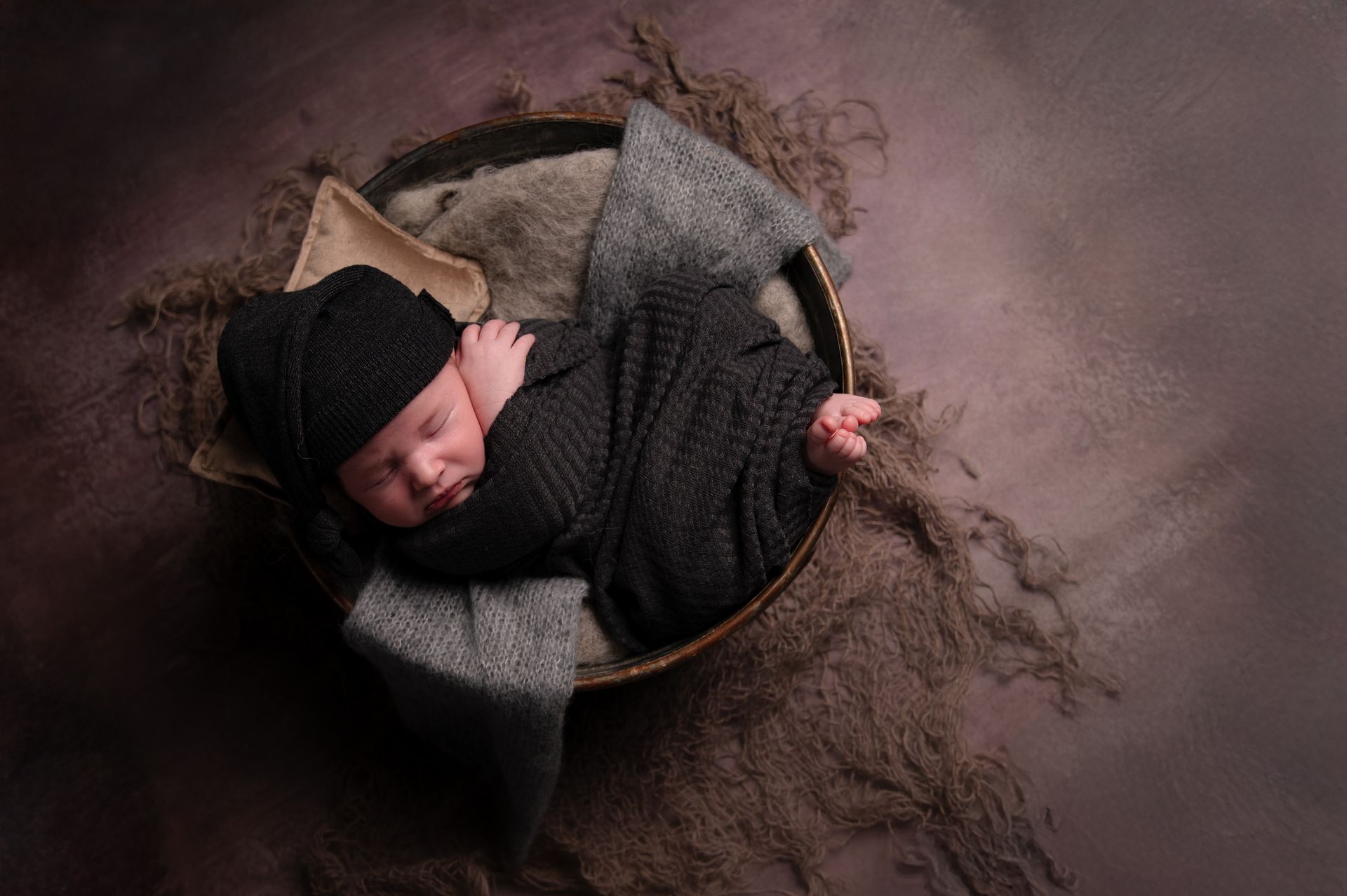 Een pasgeboren baby in een zwarte gebreide muts en omslagdoek slaapt vredig in een rustiek mandje op een bruine achtergrond.