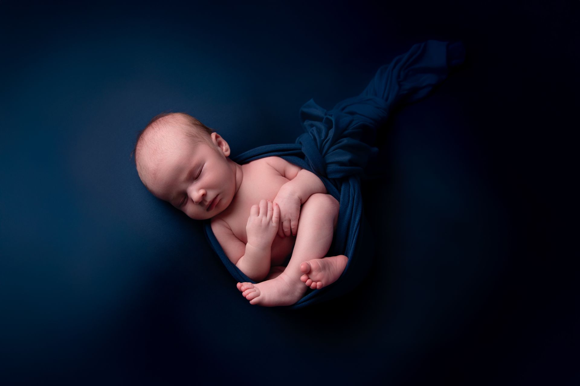 Een pasgeboren baby slaapt opgerold in een donkerblauwe doek tegen een donkerblauwe achtergrond.