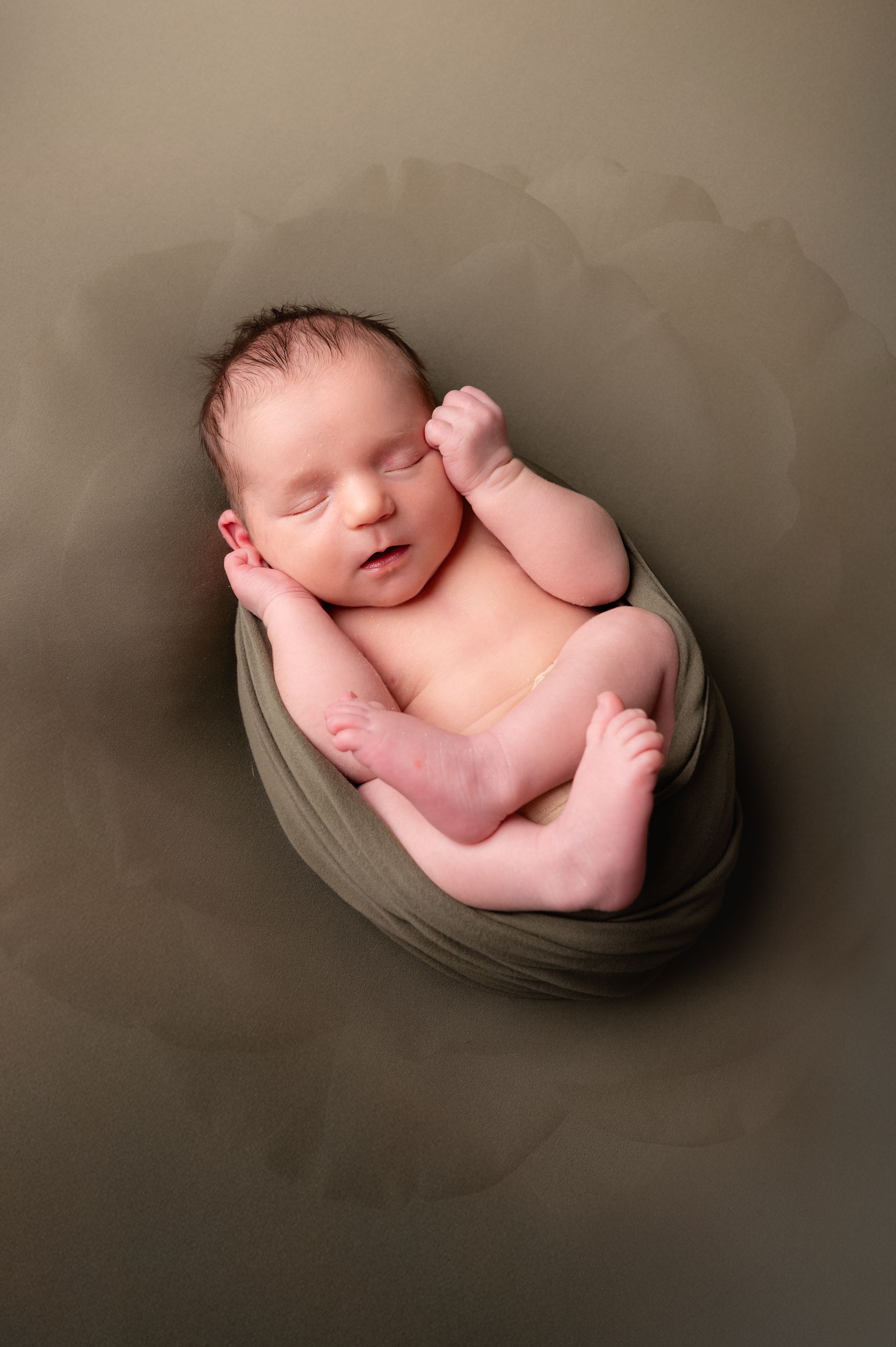 Een pasgeboren baby ligt opgerold te slapen op een groene doek, tegen een groene achtergrond.