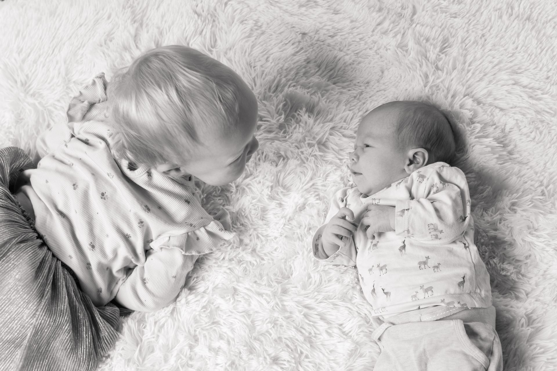 Een ouder kind kijkt naar een pasgeboren baby die op een witte, zachte ondergrond ligt. Beide kinderen dragen een lichtgekleurde pyjama.