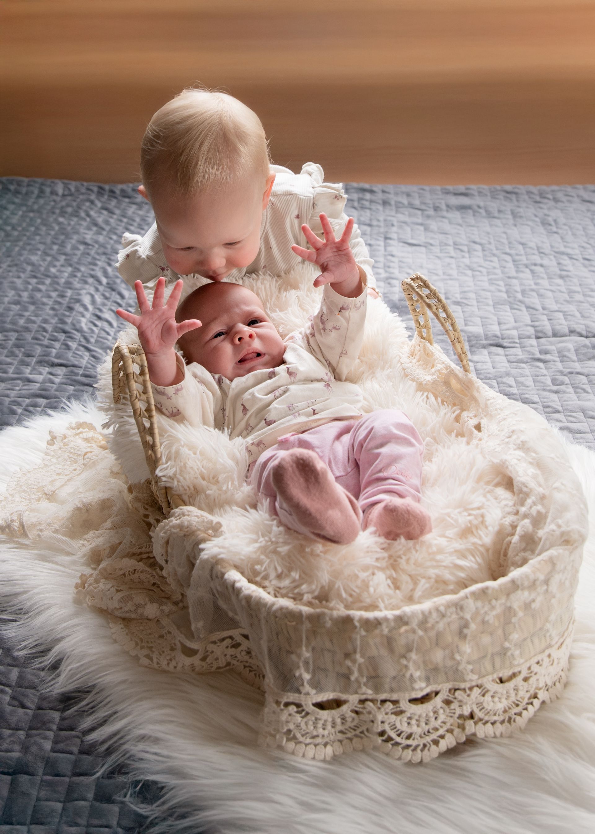 Een jong kind speelt met een baby in een mandje. De baby draagt een roze broekje en ziet er bedrukt uit. De setting is een bed met een grijze deken.