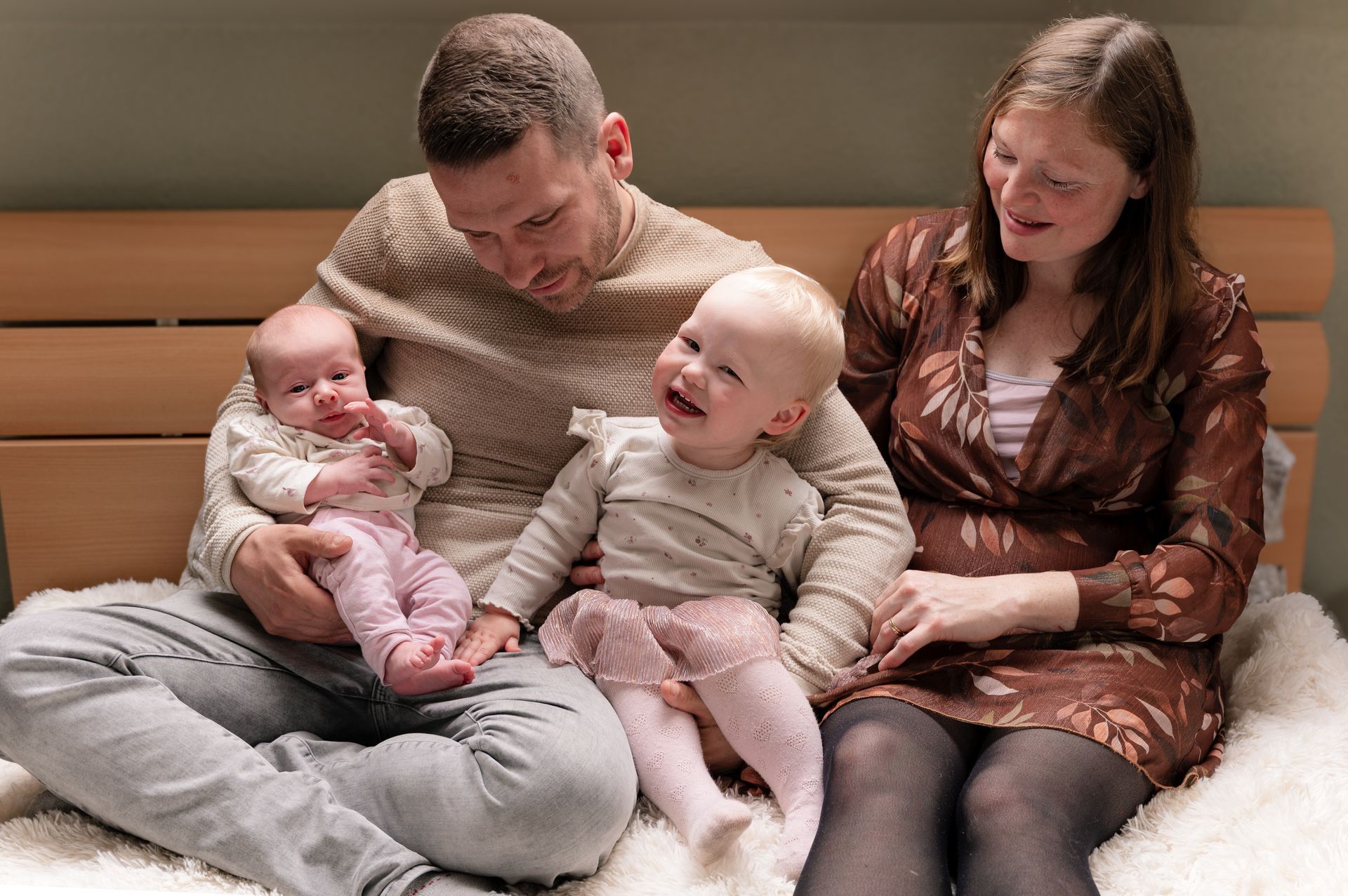 Een gezin van vier zit op een bed. De vader houdt een baby vast, de moeder zit naast een lachende peuter.