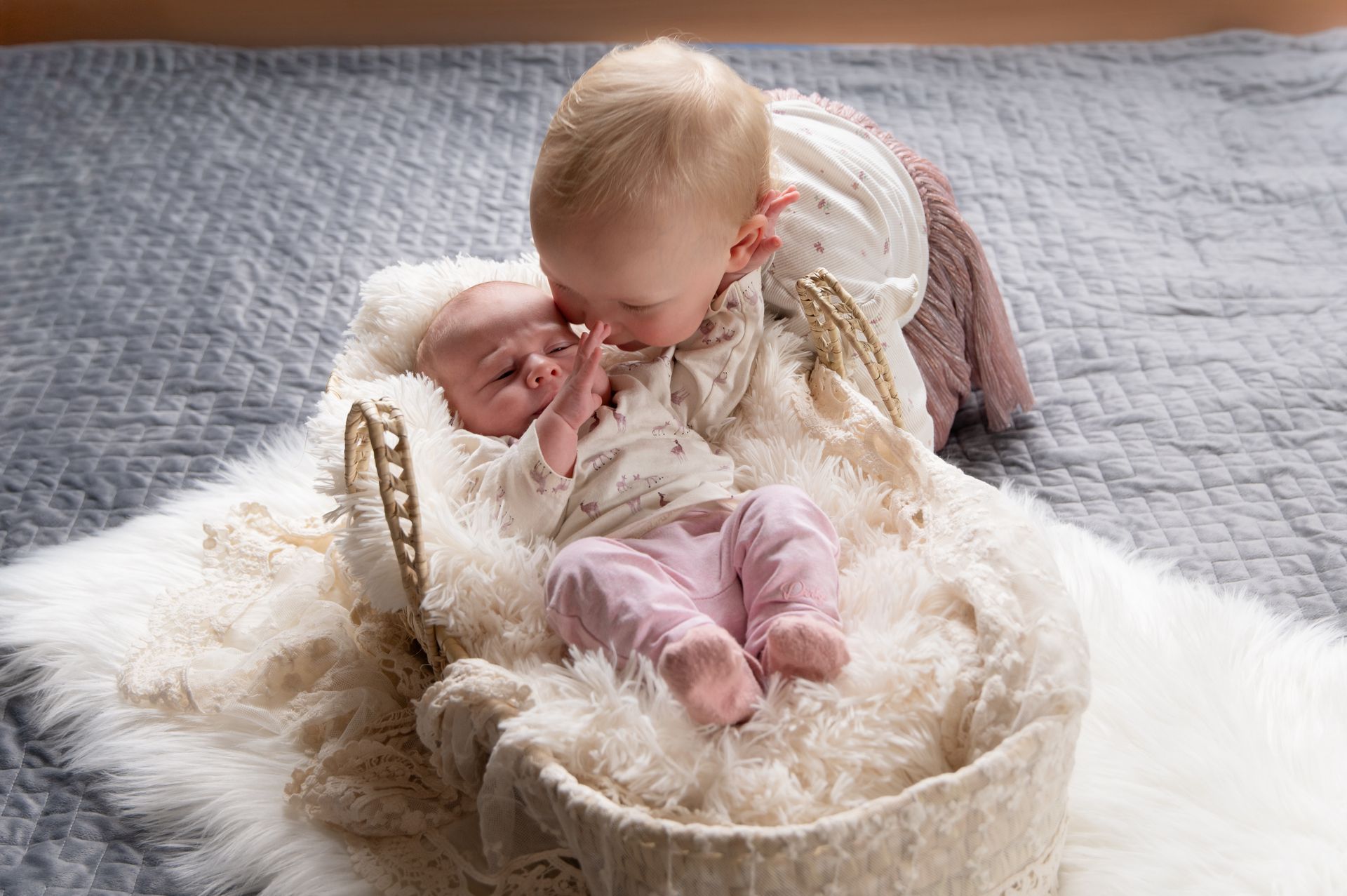 Twee baby's, één in een mandje, worden door de ander gekust. Ze liggen op een bed met zachte, wit-grijze dekens.