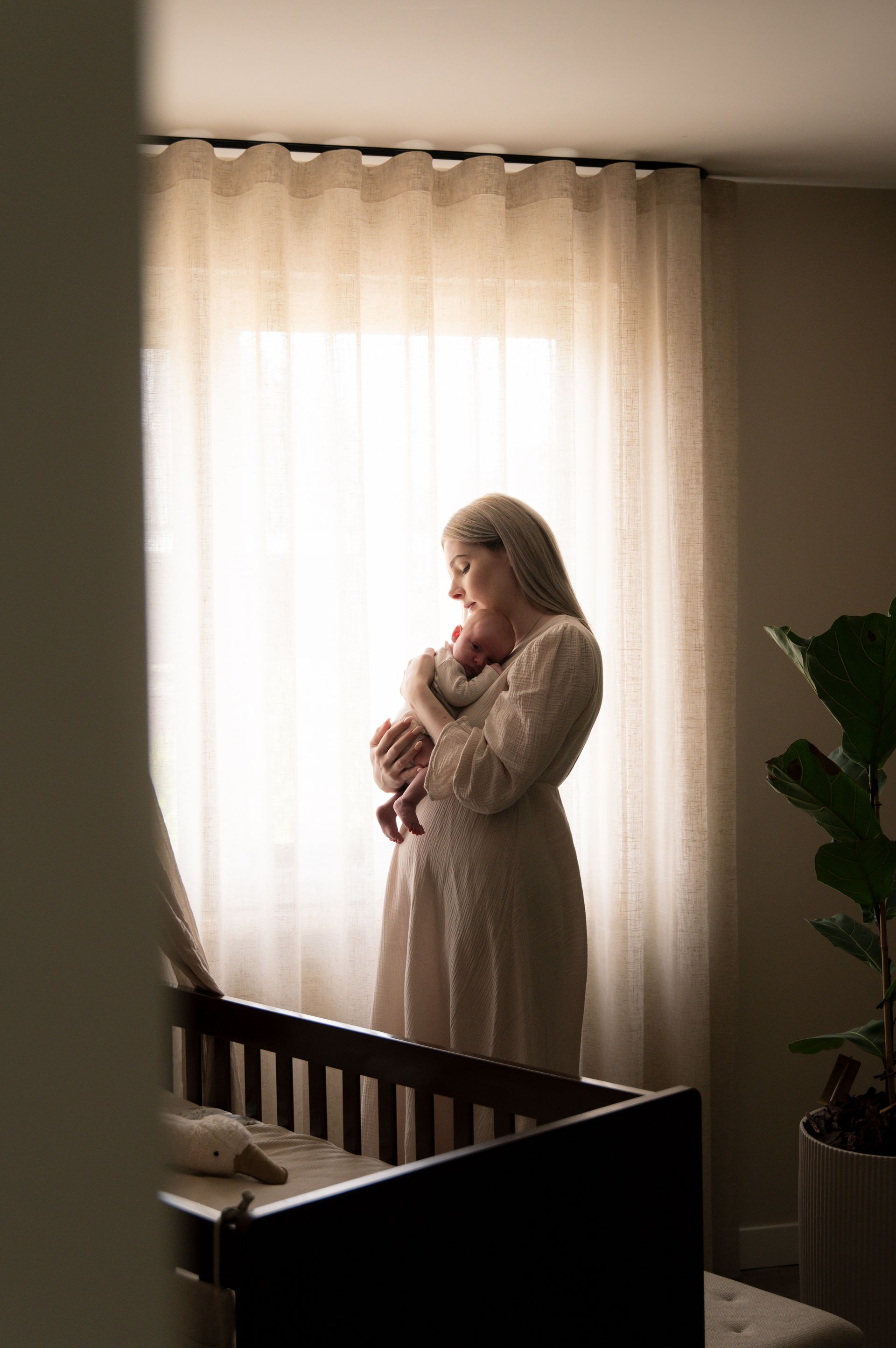 Een vrouw in een lange jurk houdt een pasgeboren baby vast bij een raam in een kinderkamer, met een wiegje zichtbaar.