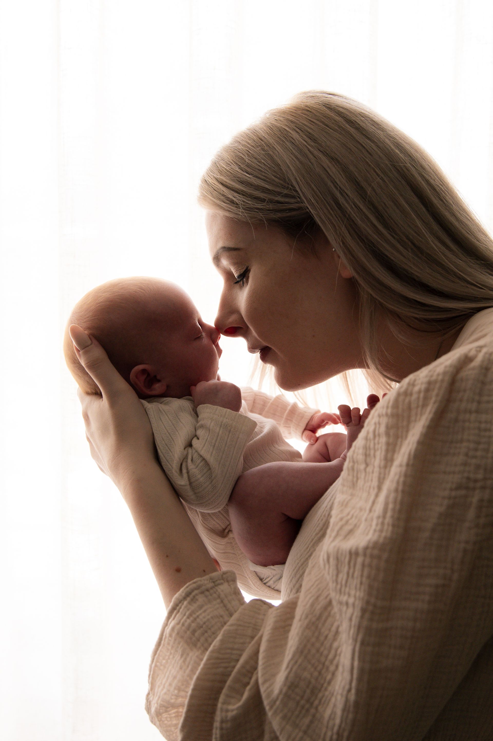 Moeder met een pasgeboren baby, met de neusjes tegen elkaar, voor een fel verlicht raam. De moeder heeft blond haar en draagt een lichtgekleurd gewaad.