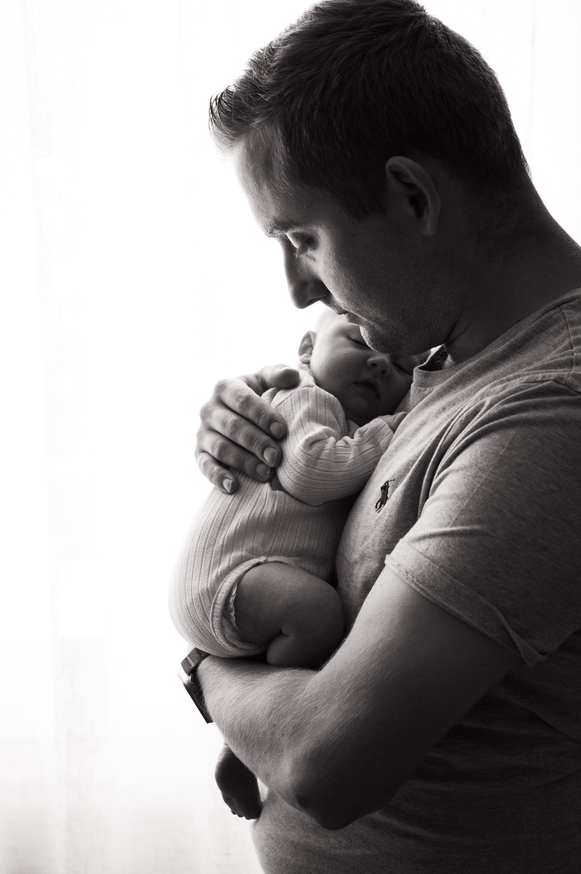 Zwart-witfoto van een man die een slapende baby tegen zijn borst houdt. De man kijkt met een zachte blik naar beneden.