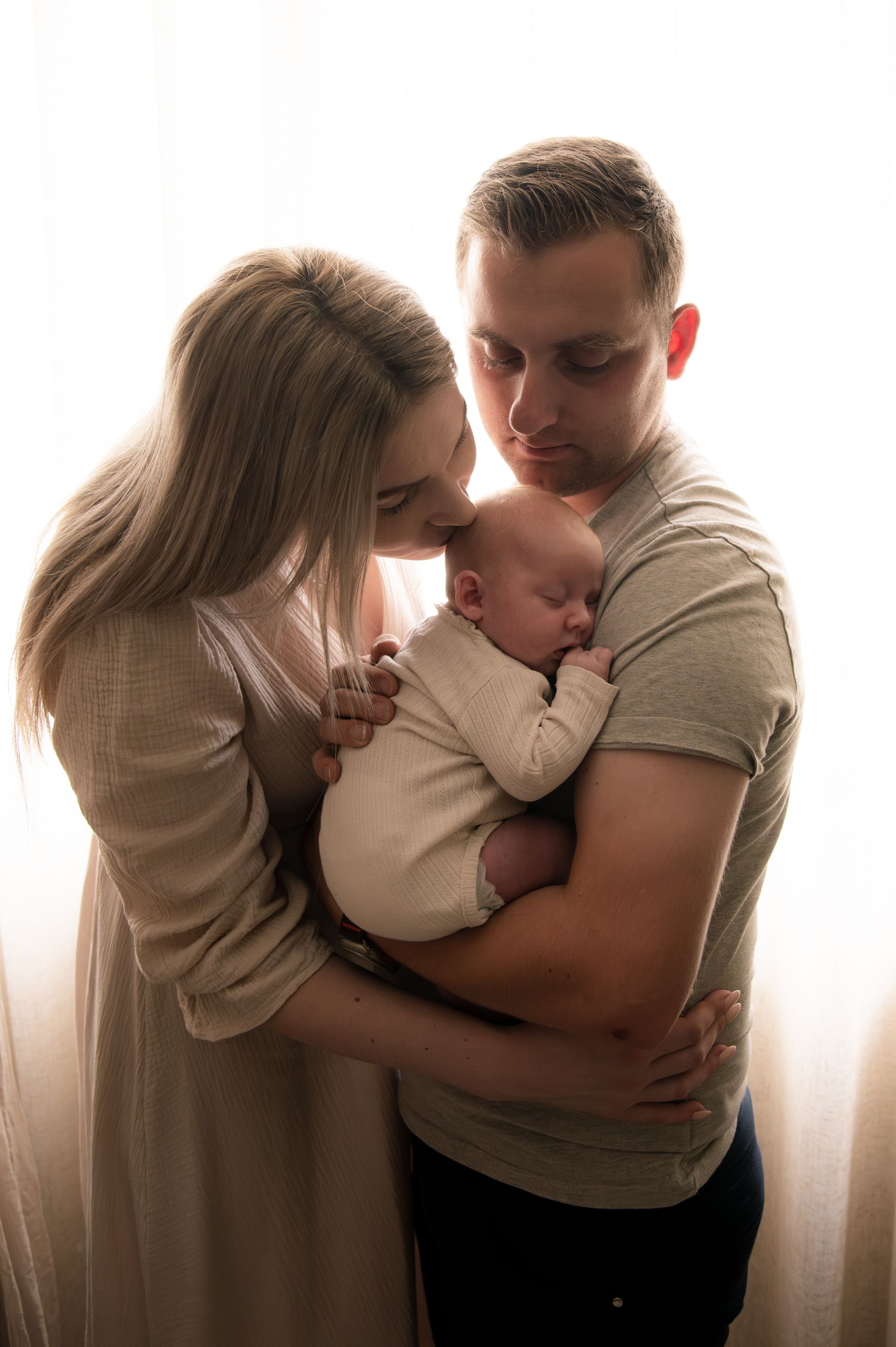 Een pasgeboren baby slaapt in de armen van zijn vader, terwijl de moeder hem een kus op zijn hoofdje geeft. Het gezin wordt van achteren verlicht door een helder raam.