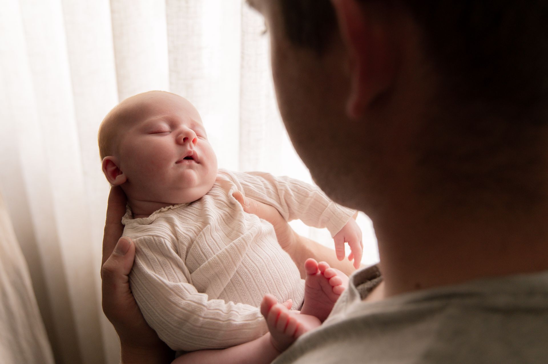 Een persoon houdt een slapende baby vast bij een raam. De baby is bleek, heeft gesloten ogen en de persoon is onscherp.