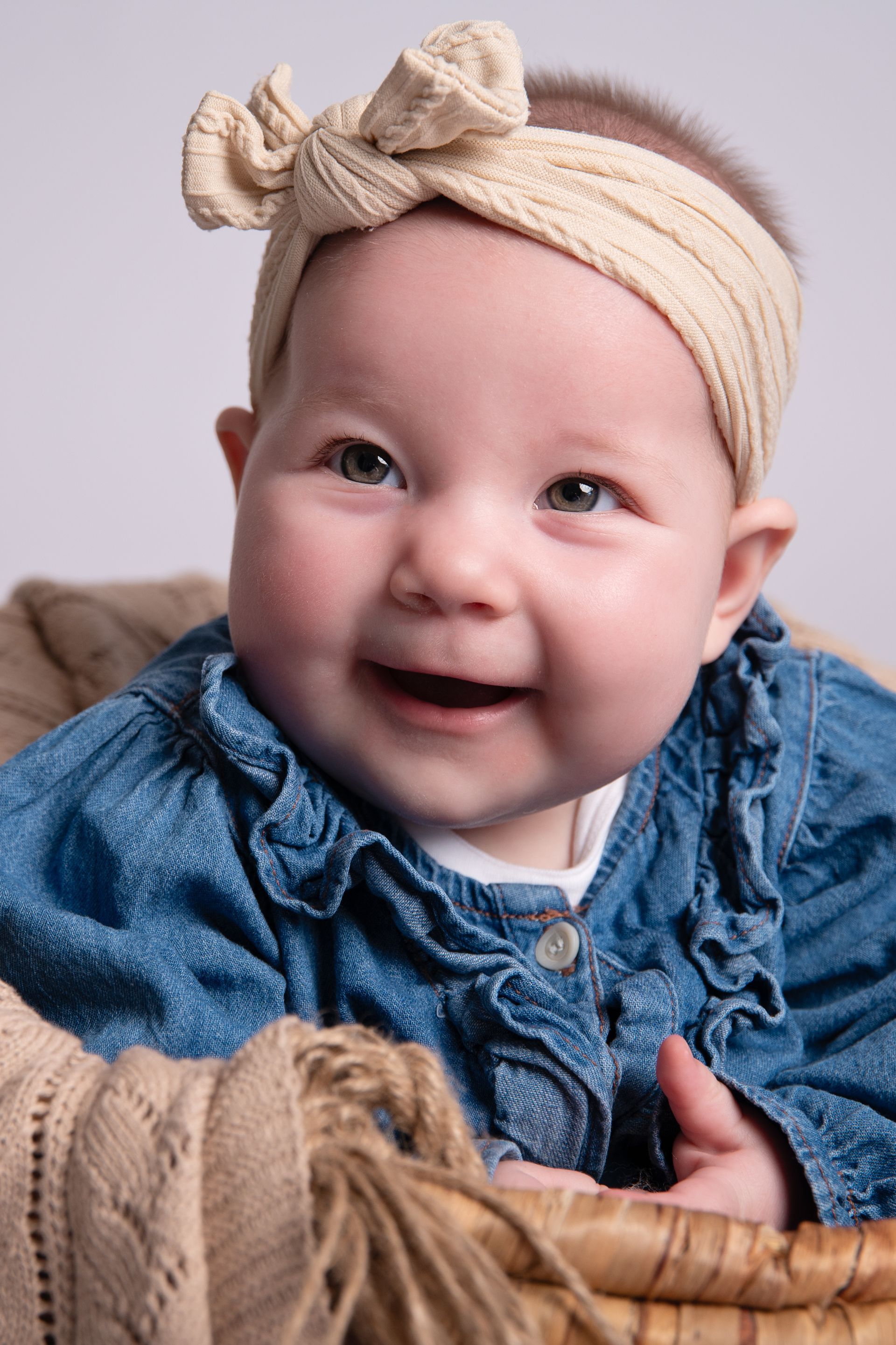 Een babymeisje met een haarband zit in een mandje en lacht.