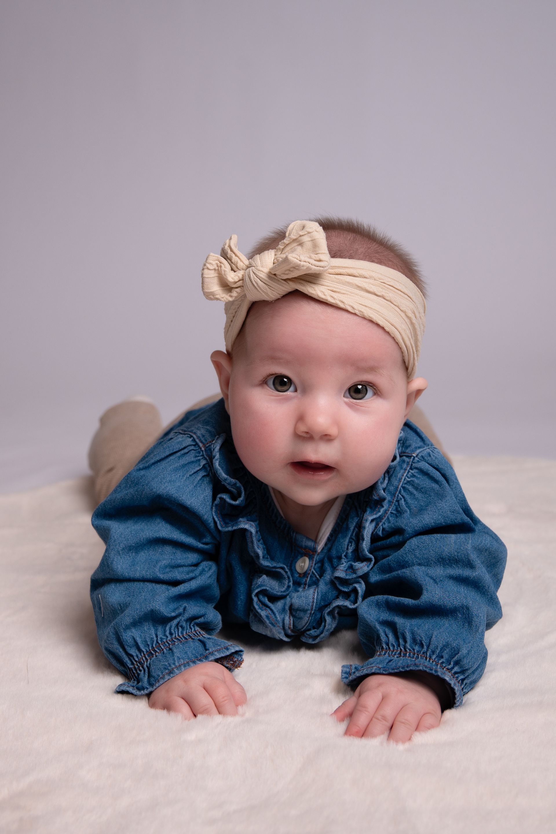 Een babymeisje draagt ​​een spijkerblouse en een haarband en ligt op haar buik op een deken.