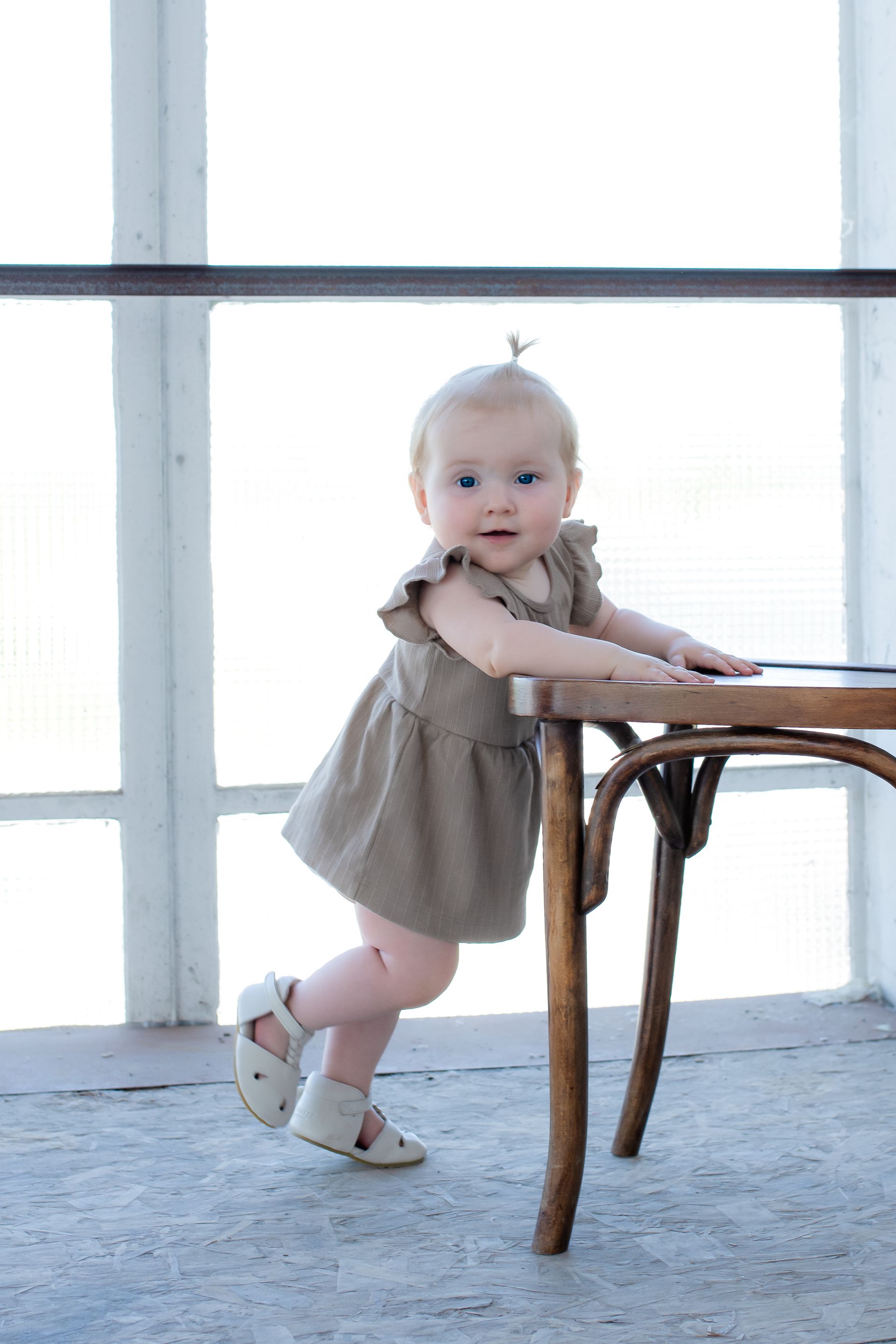 Een babymeisje in een jurk staat naast een stoel.