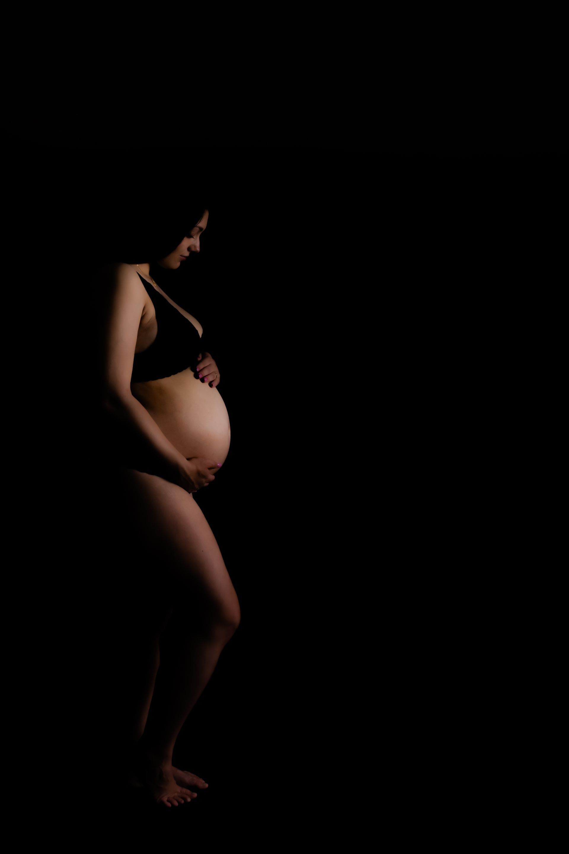 Een zwangere vrouw staat in het donker met haar handen op haar buik.