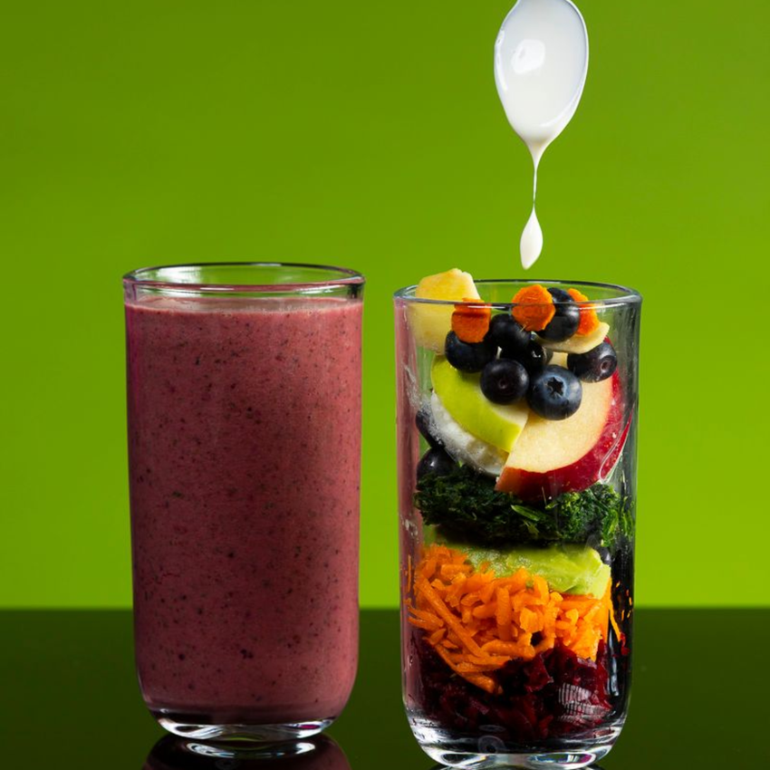Glass of purple smoothie next to a glass layered with fruits and vegetables, white liquid drips on top.