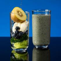 Glass of ingredients next to a finished smoothie. Ingredients: kiwi, banana, blueberries, greens. Blue background.