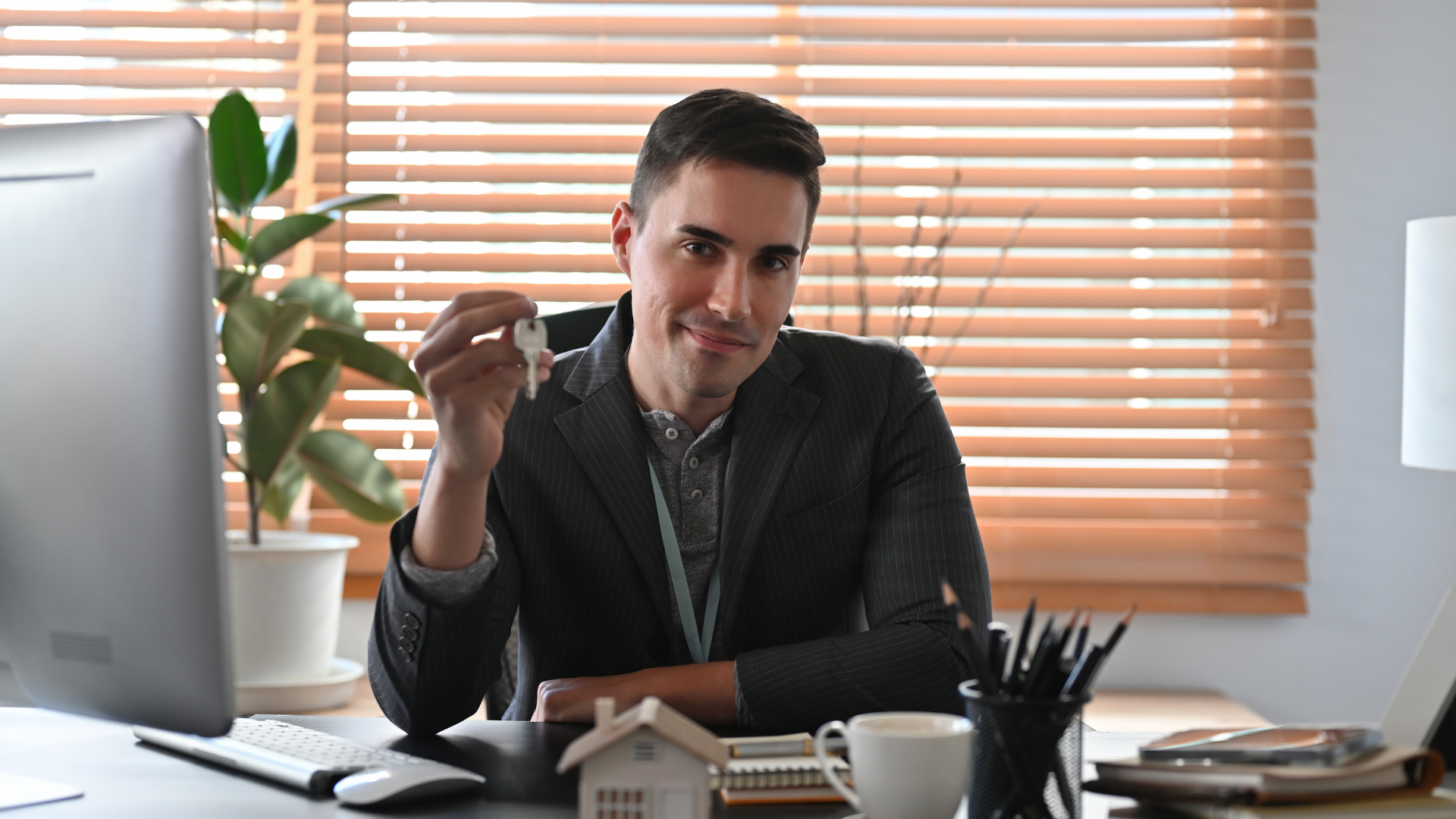 Man holding a key and small house model smiles at desk in office setting.