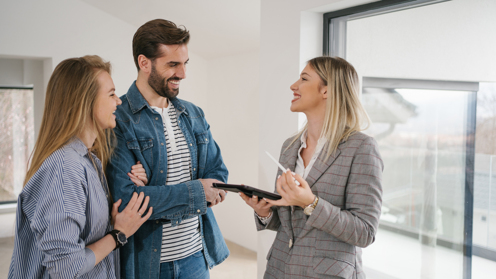 A real estate agent showing a house to a smiling couple, standing by a window.