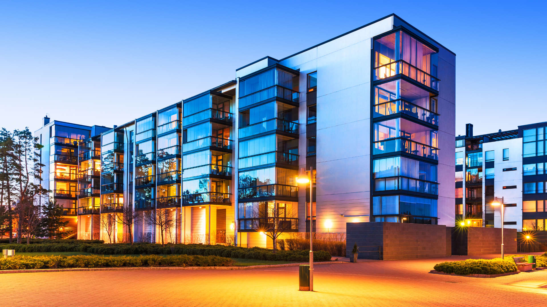 Modern multi-story apartment building with glass balconies and lit windows at dusk.