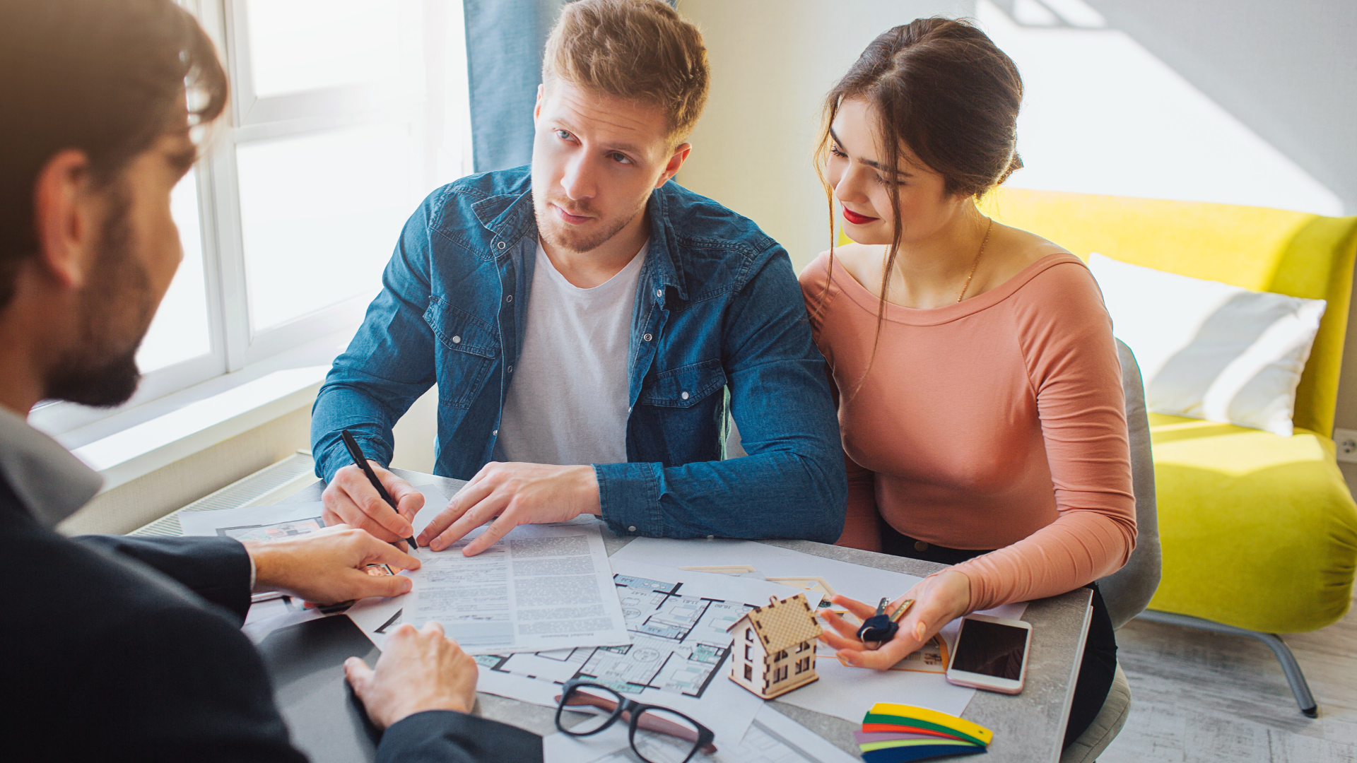Couple signing a contract at a table with a realtor, house model, and phone.