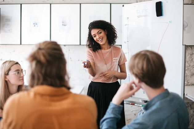 Woman in a meeting, gesturing, smiling at whiteboard, with three seated colleagues in a bright office.