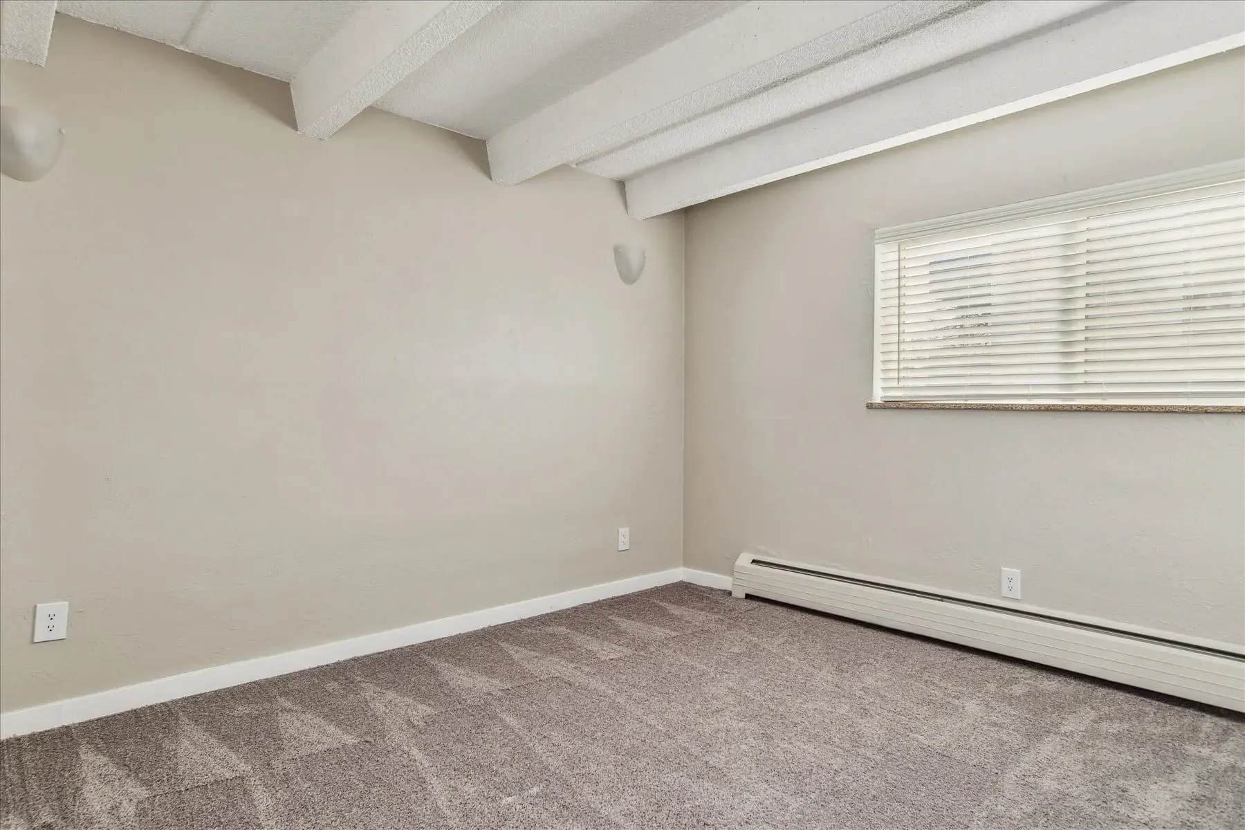 Empty apartment bedroom with beige walls and carpet. Features a window with blinds and a radiator.