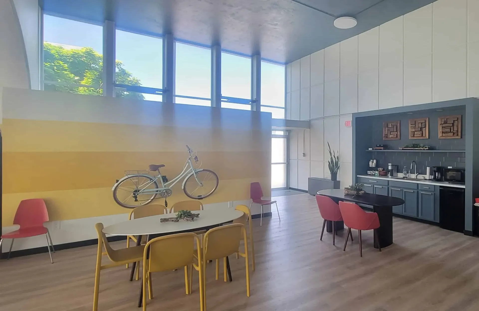 Modern community lounge area with seating, a kitchenette, and large windows overlooking greenery.