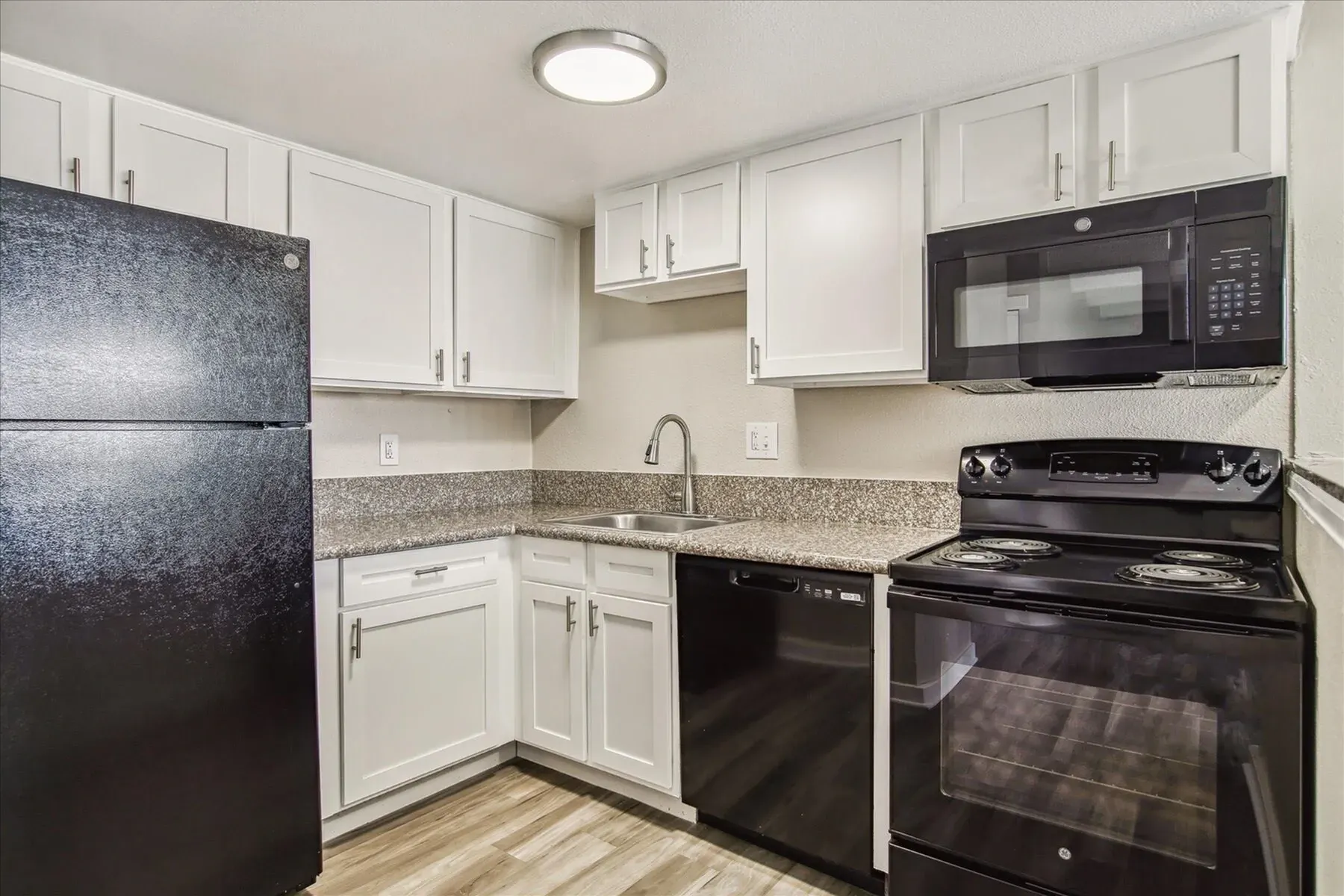 A clean, modern kitchen with white cabinets, granite countertops, and black appliances, including a refrigerator, dishwasher, oven, and microwave.