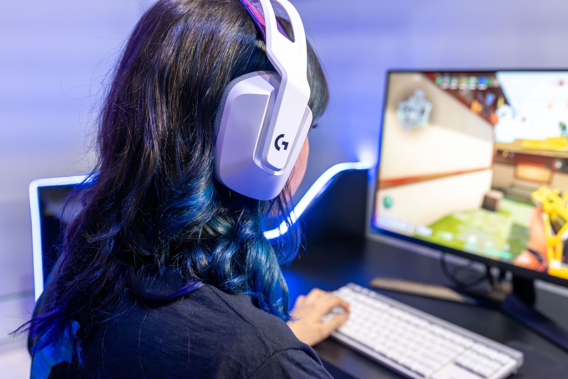 Person wearing white headphones gaming at a desktop computer in a blue-lit room