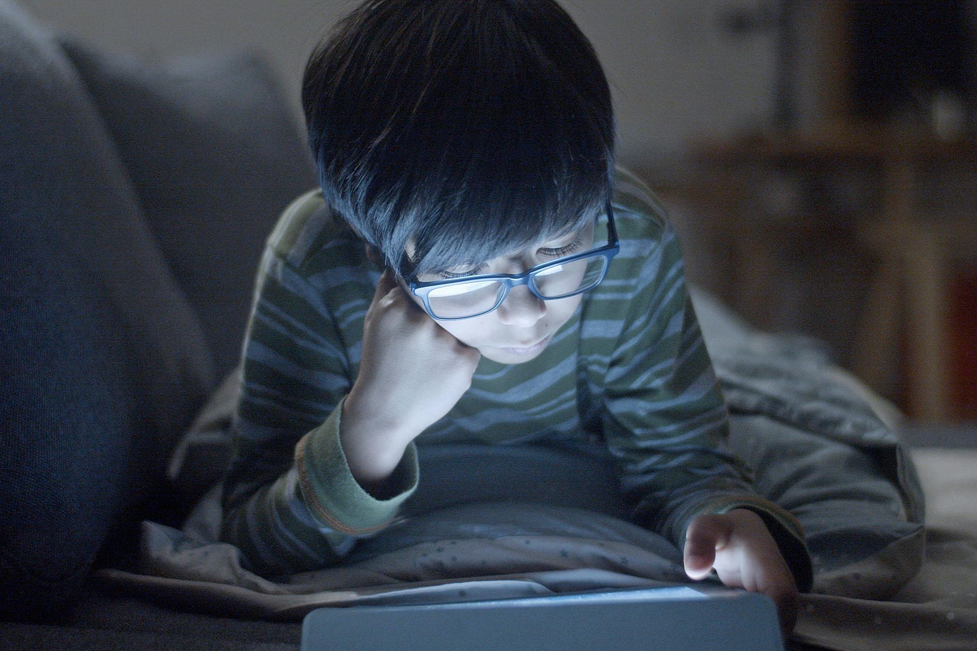 Child in striped shirt reading a glowing tablet on a couch in the dark