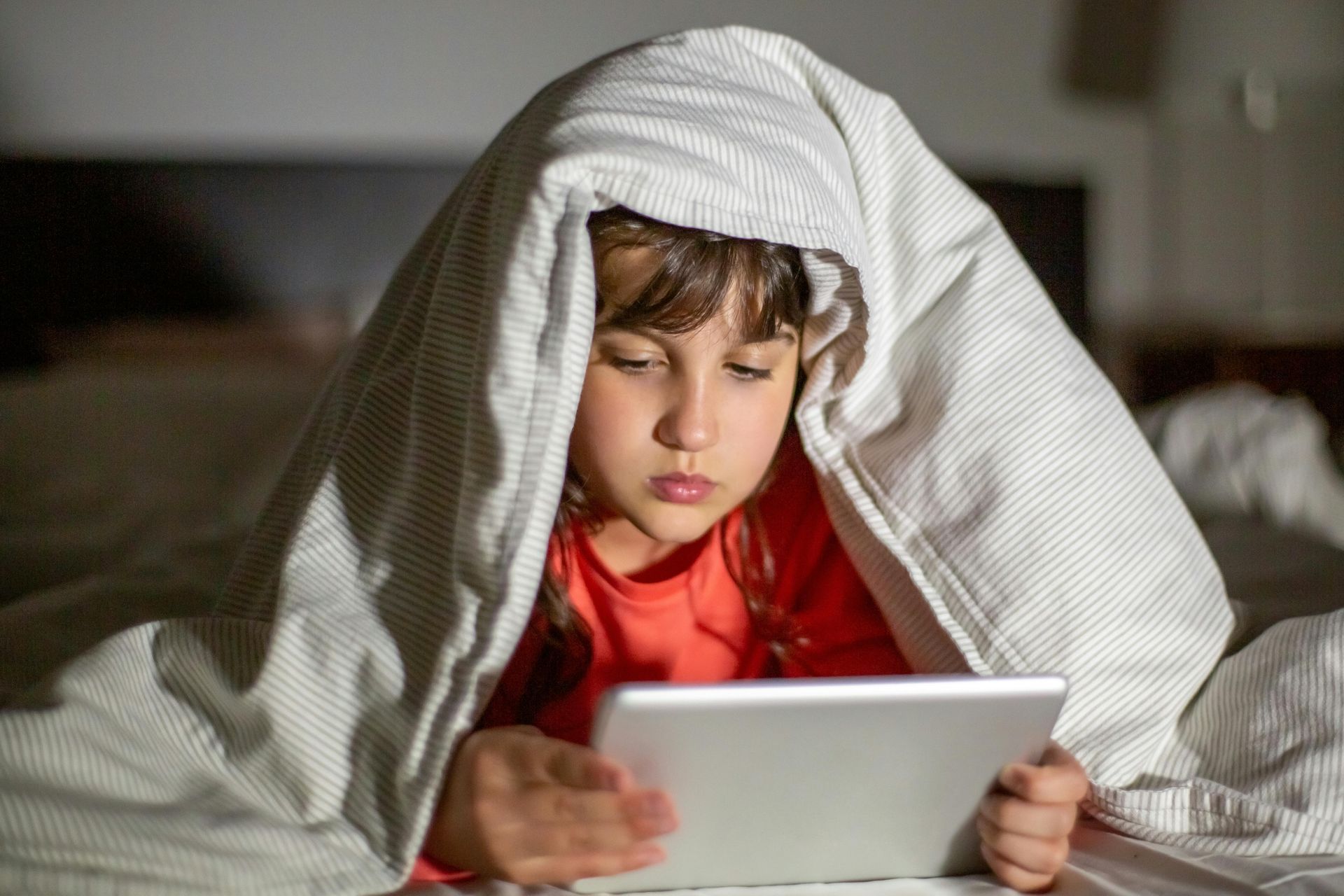 Child under a gray blanket using a tablet, focused in a dim room