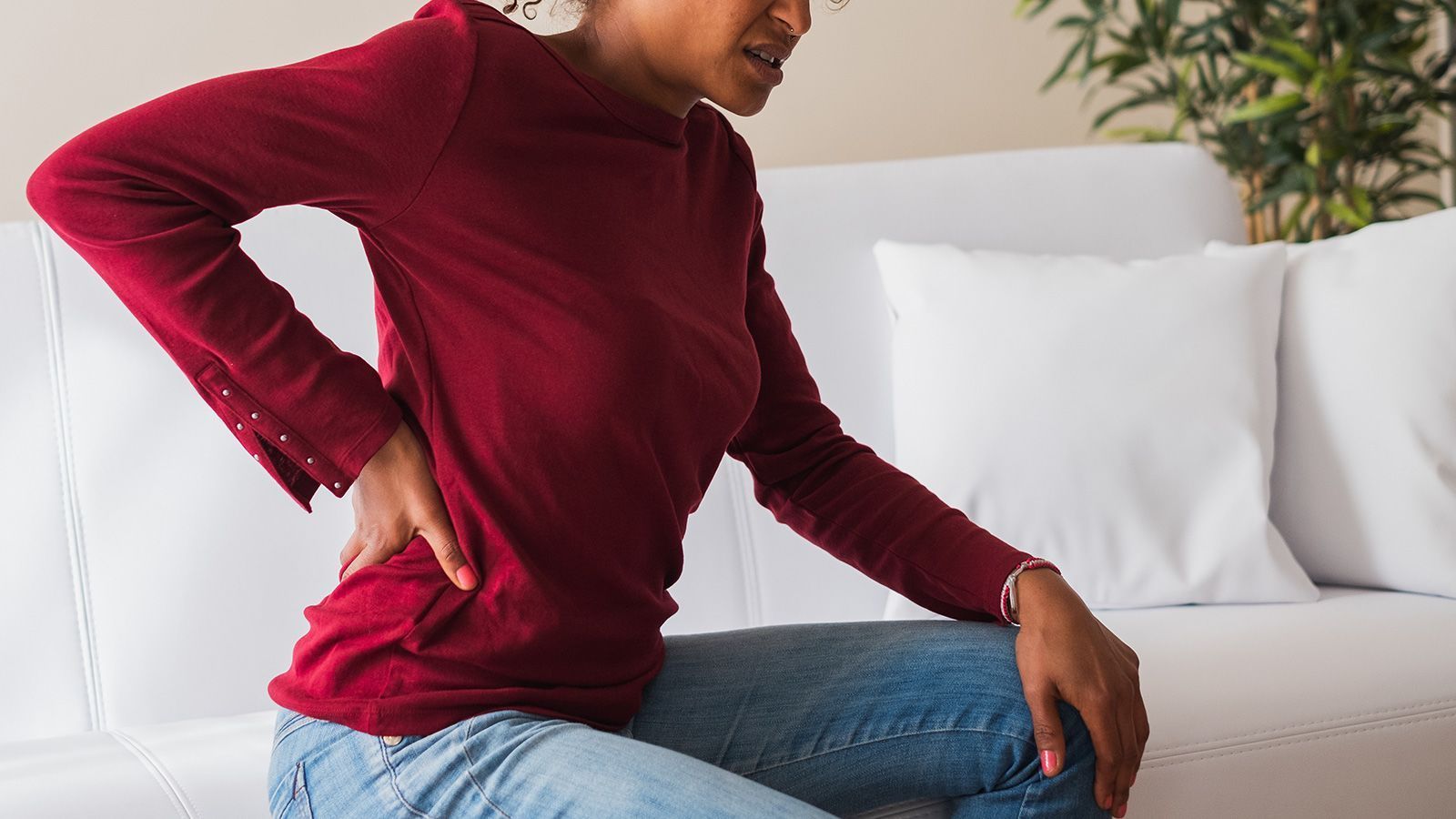A woman is sitting on a couch holding her back in pain.