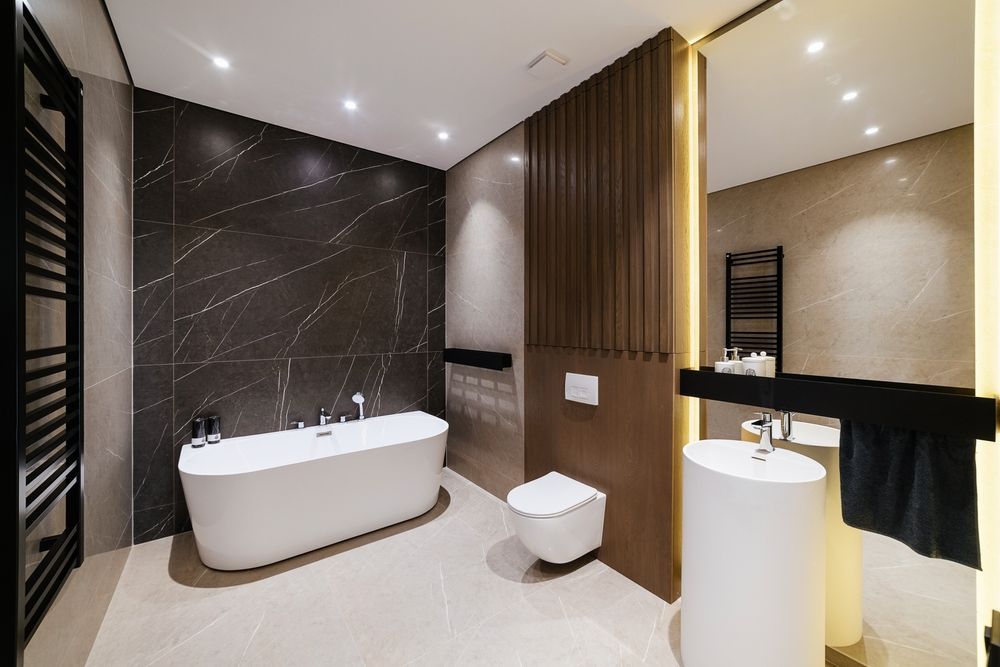 Modern bathroom with a freestanding white tub, wall-mounted toilet, and pedestal sink against dark stone and wood panels.