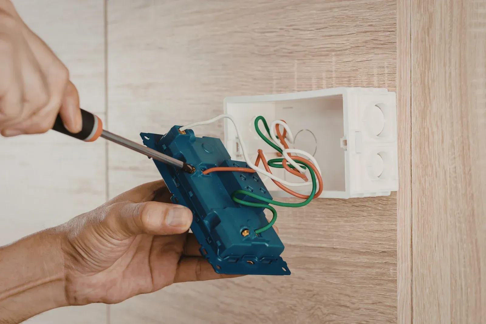 Person using screwdriver to install electrical switch into wall box; exposed wires visible.