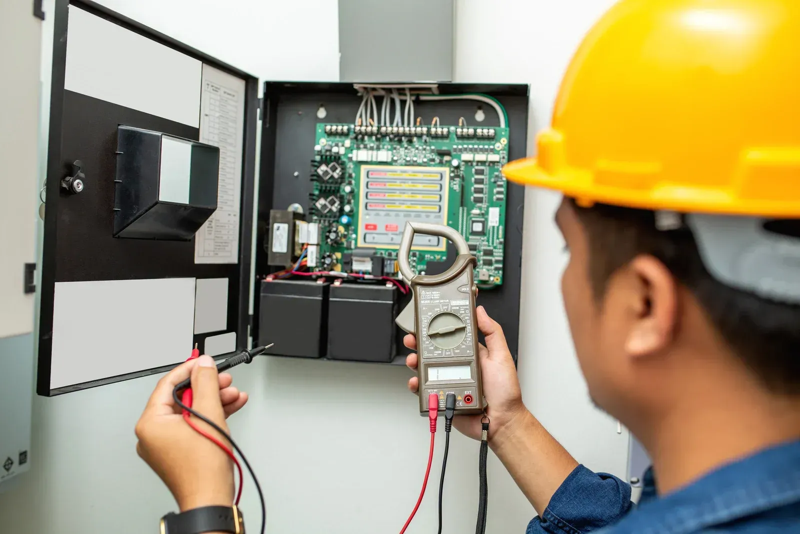 Electrician in yellow hard hat using a multimeter to inspect an electrical panel with circuit boards and batteries.