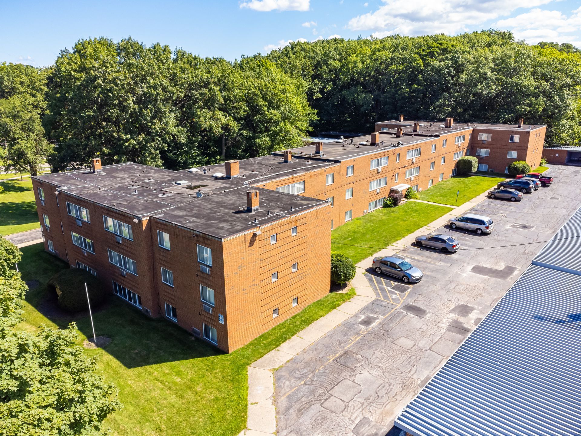 Red brick apartment building with parked cars on a sunny day, surrounded by green trees.