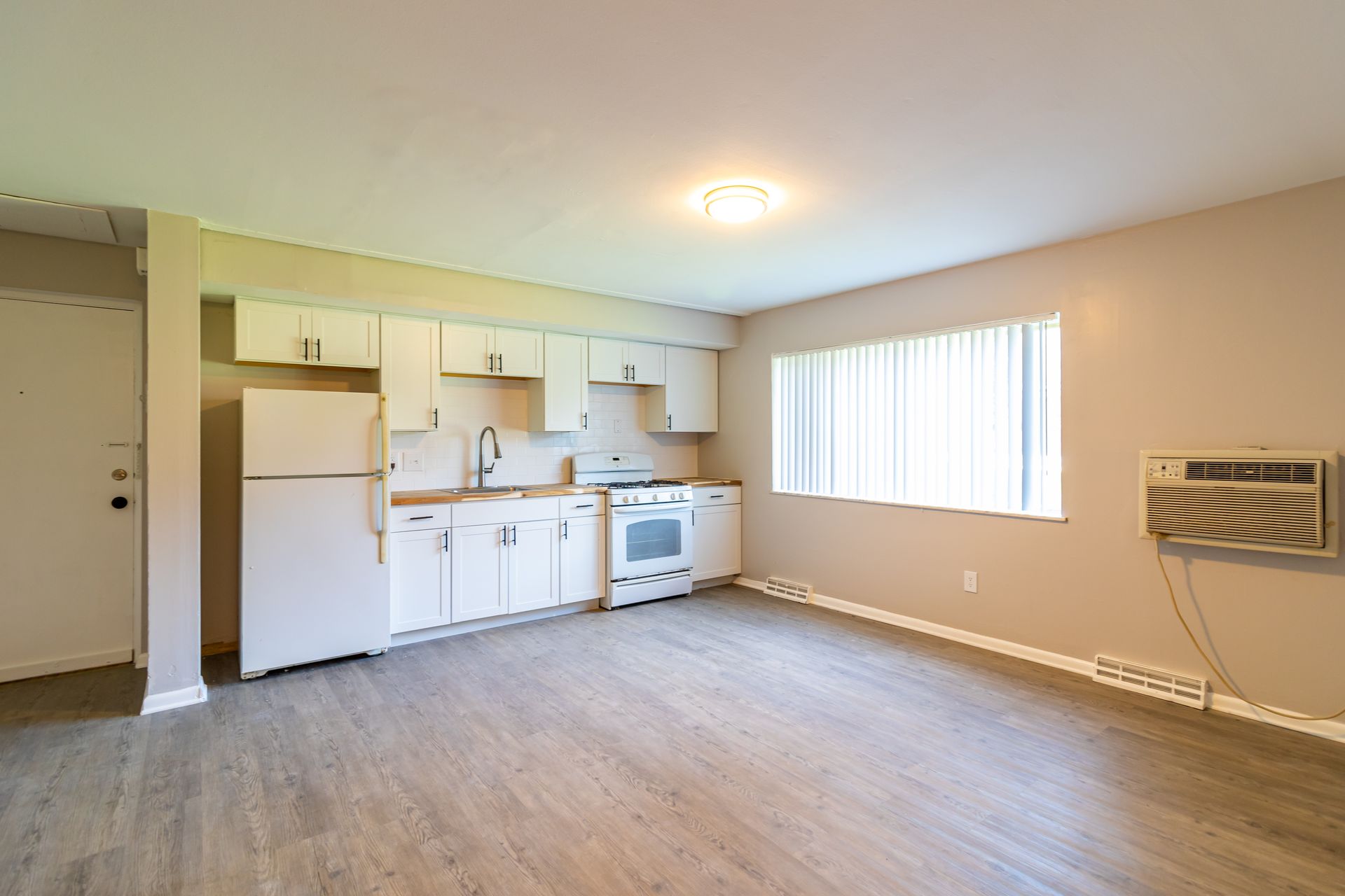 Open-concept kitchen and living area with white cabinets, appliances, and tan walls. Window with blinds on right.