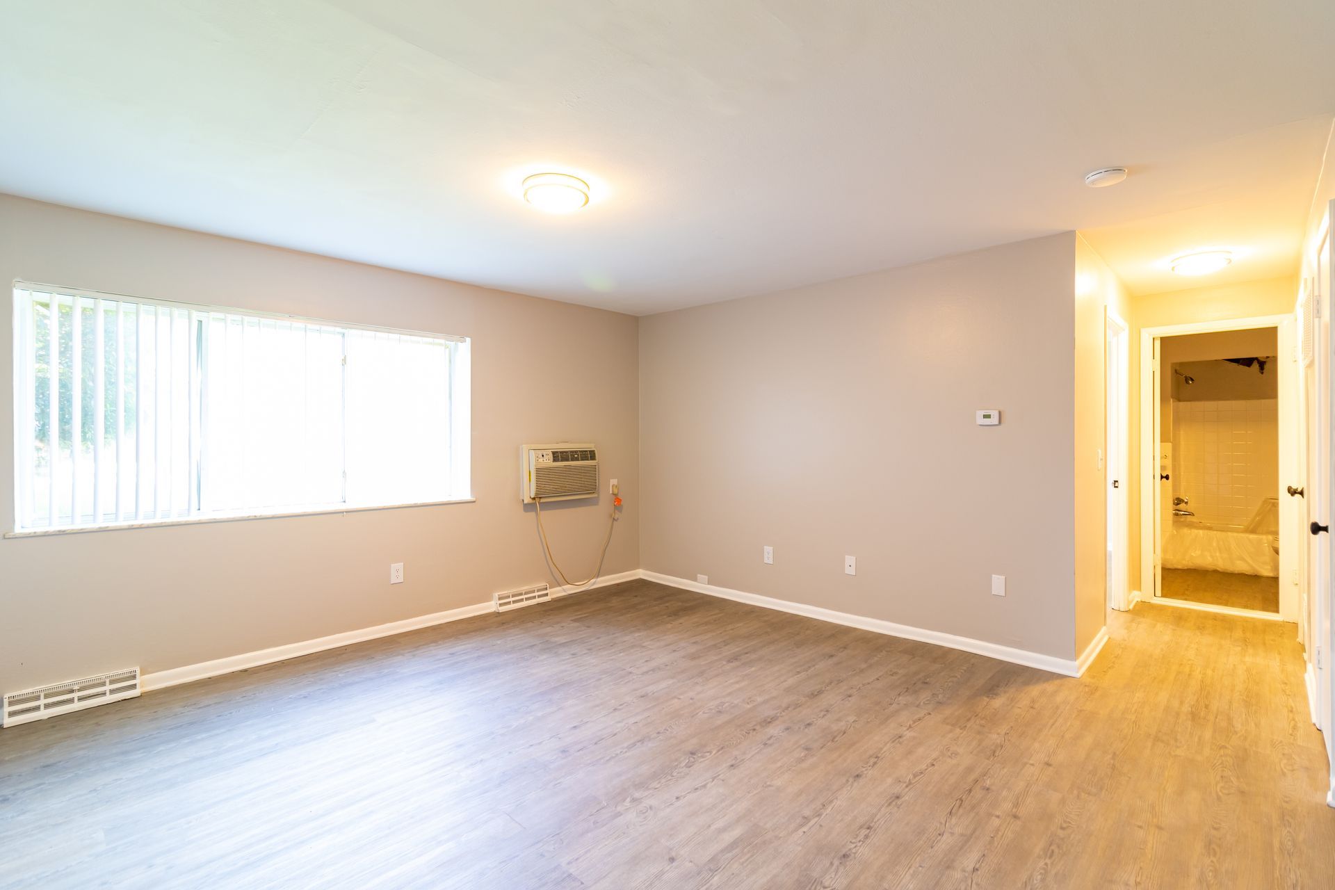 Empty apartment interior: light walls, wood-look floor, window with blinds, AC unit, hallway to a bathroom.