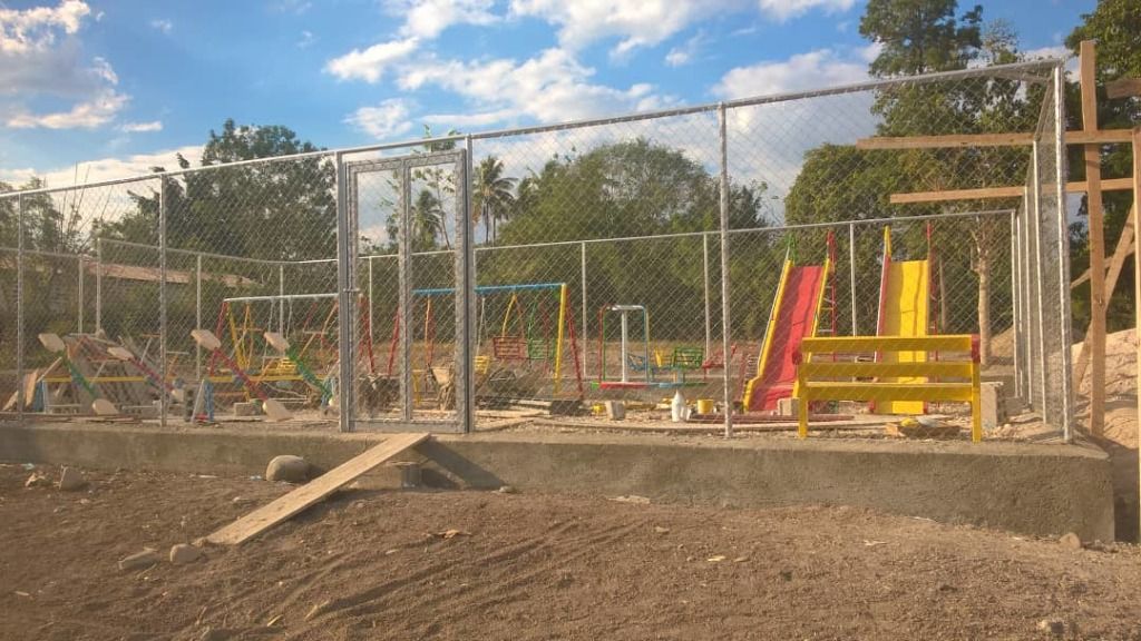 A fenced playground with colorful slides and swings sits on sandy ground.