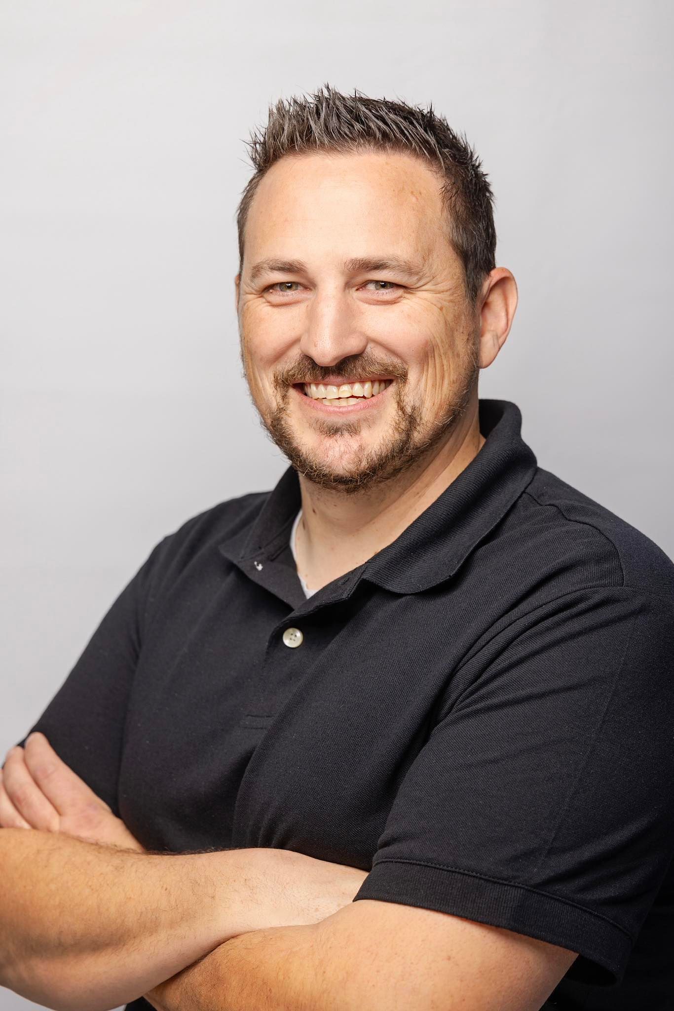A smiling man with short brown hair and a dark polo shirt stands with his arms crossed against a plain white background.