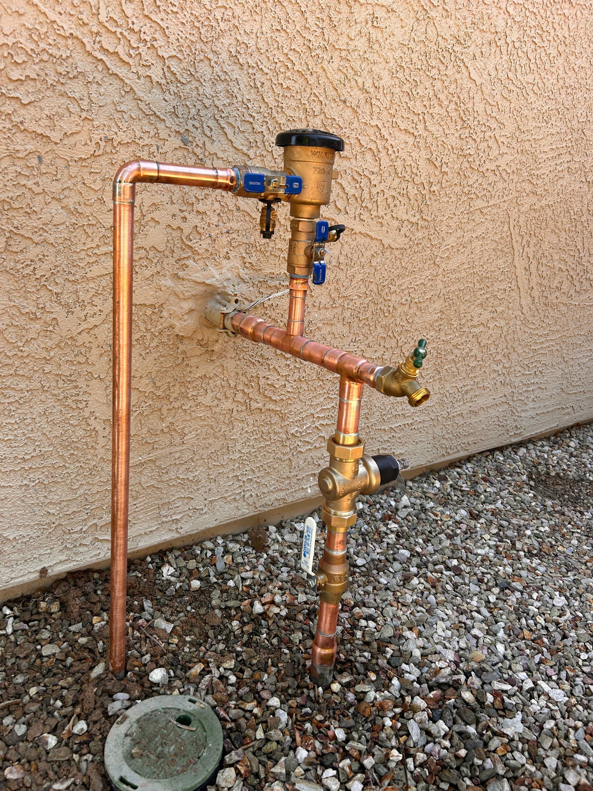 A brass backflow prevention assembly next to a water spigot, with a white arrow pointing to the main water shut-off valve.