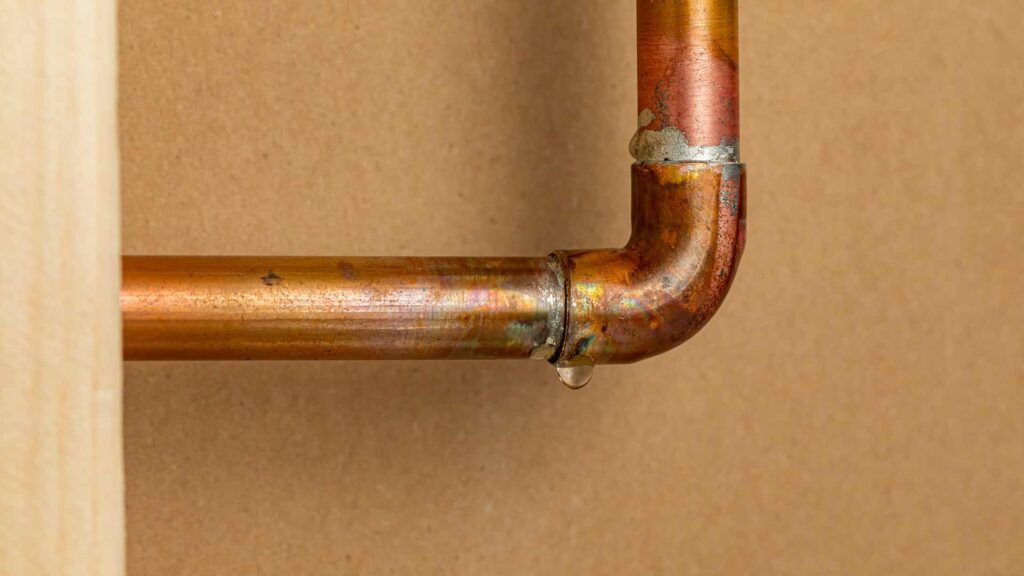 A leaking copper pipe joint with a visible water droplet, attached to a wooden wall.