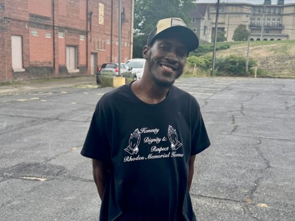 Man in a black t-shirt and cap smiles outdoors, in front of buildings and parked cars.