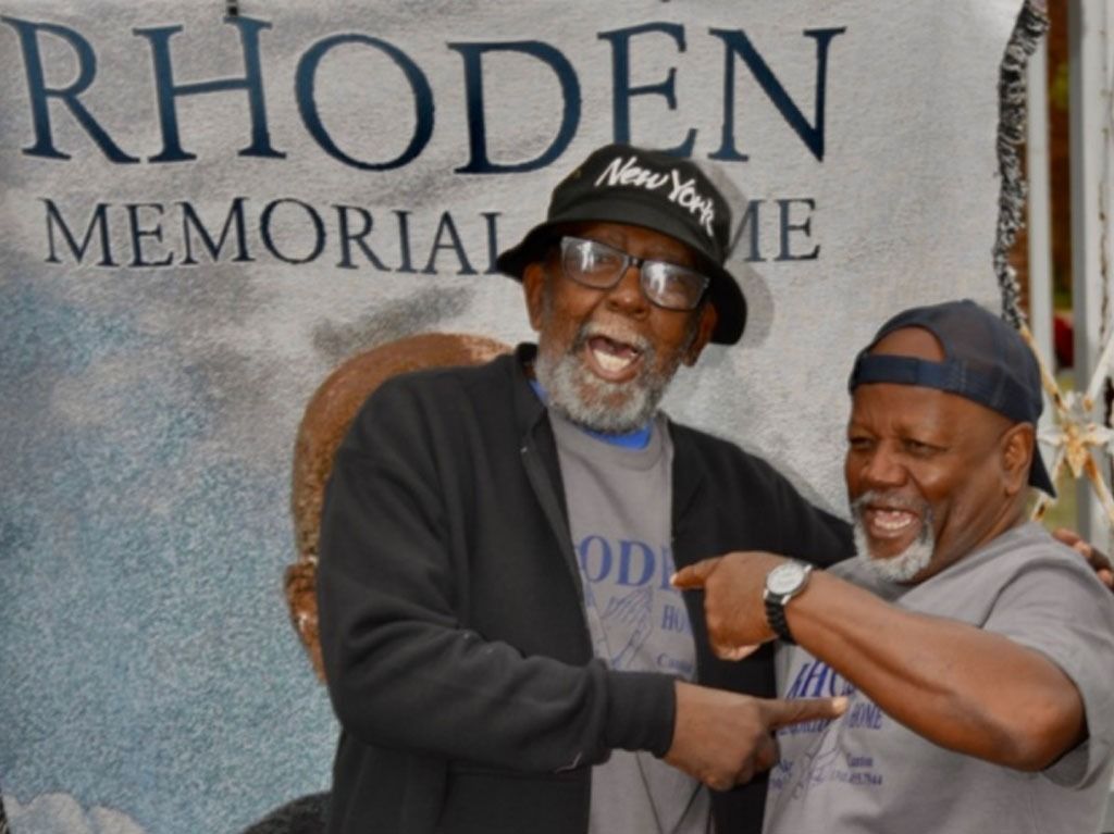 Two older Black men smile and point. One wears a